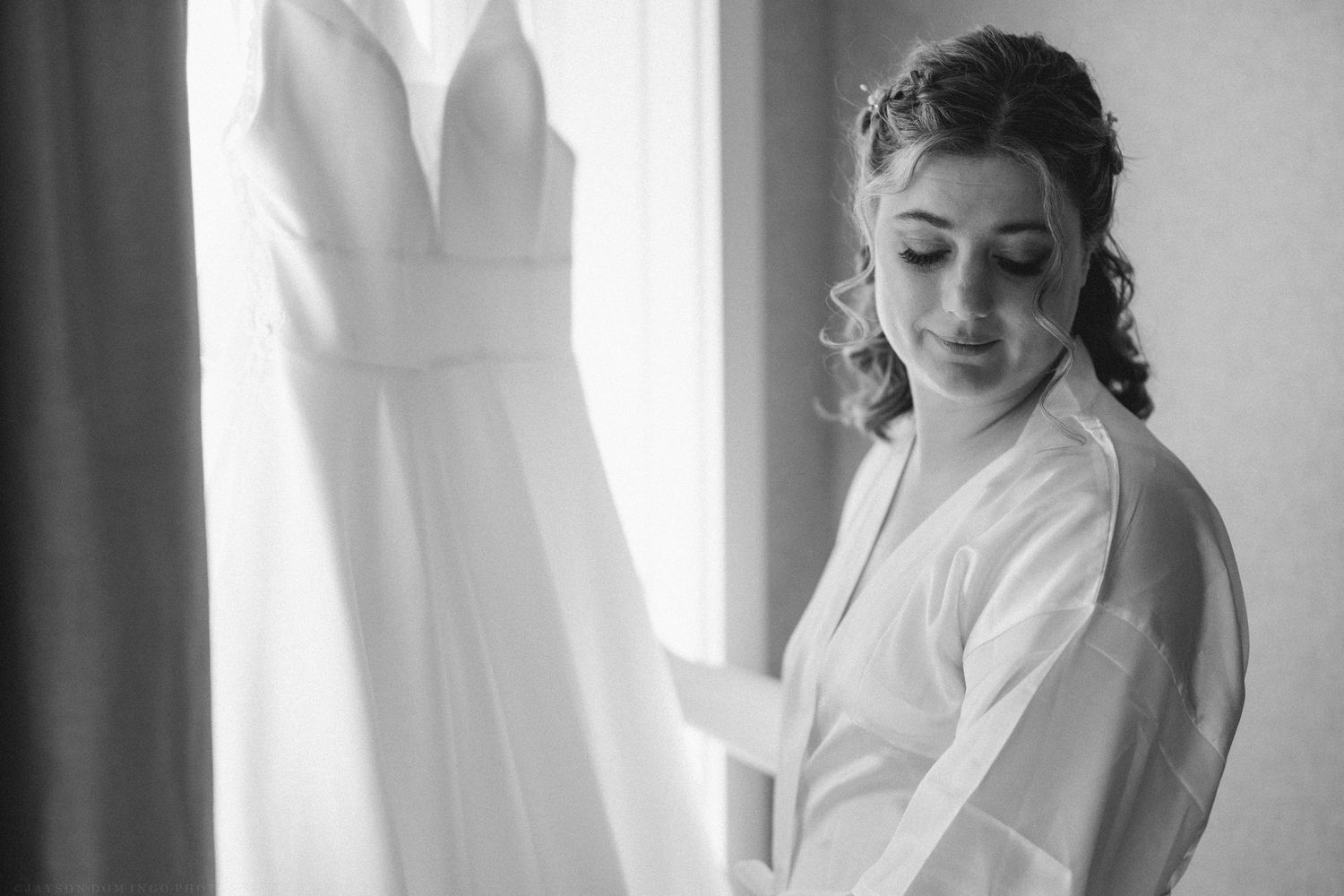 Black and white photo of a bride in a robe admiring her elegant wedding dress hanging by a window.