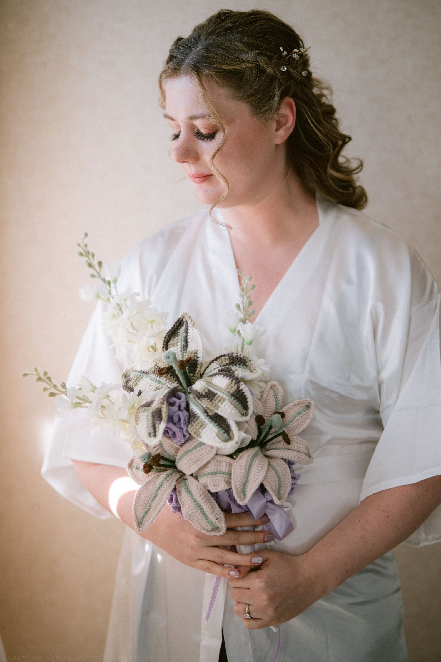 Bride in white robe holding unique handcrafted lily wedding bouquet with purple accents.