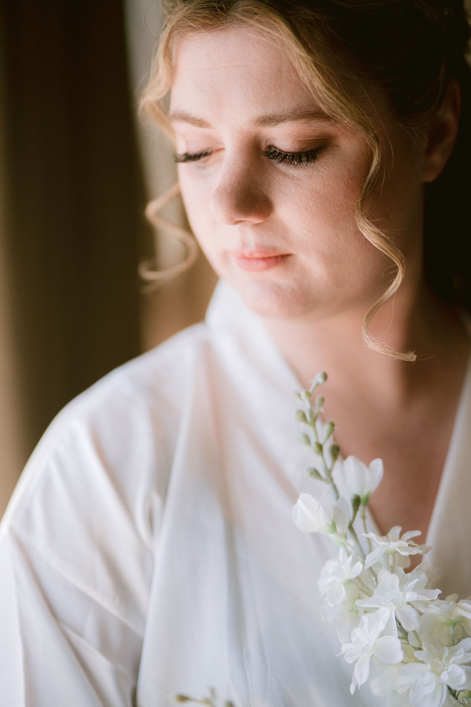 A bride in a white robe holds delicate white flowers, eyes closed, in soft natural light.