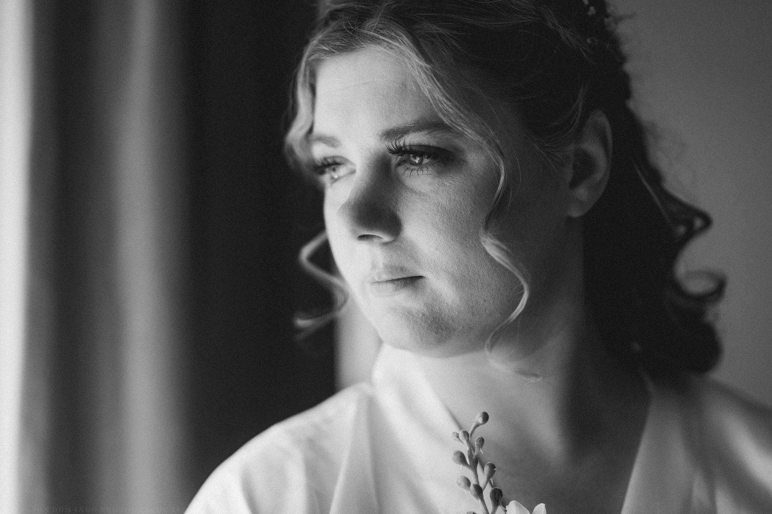 Black and white portrait of a bride in a white robe holding flowers, gazing thoughtfully out a window.