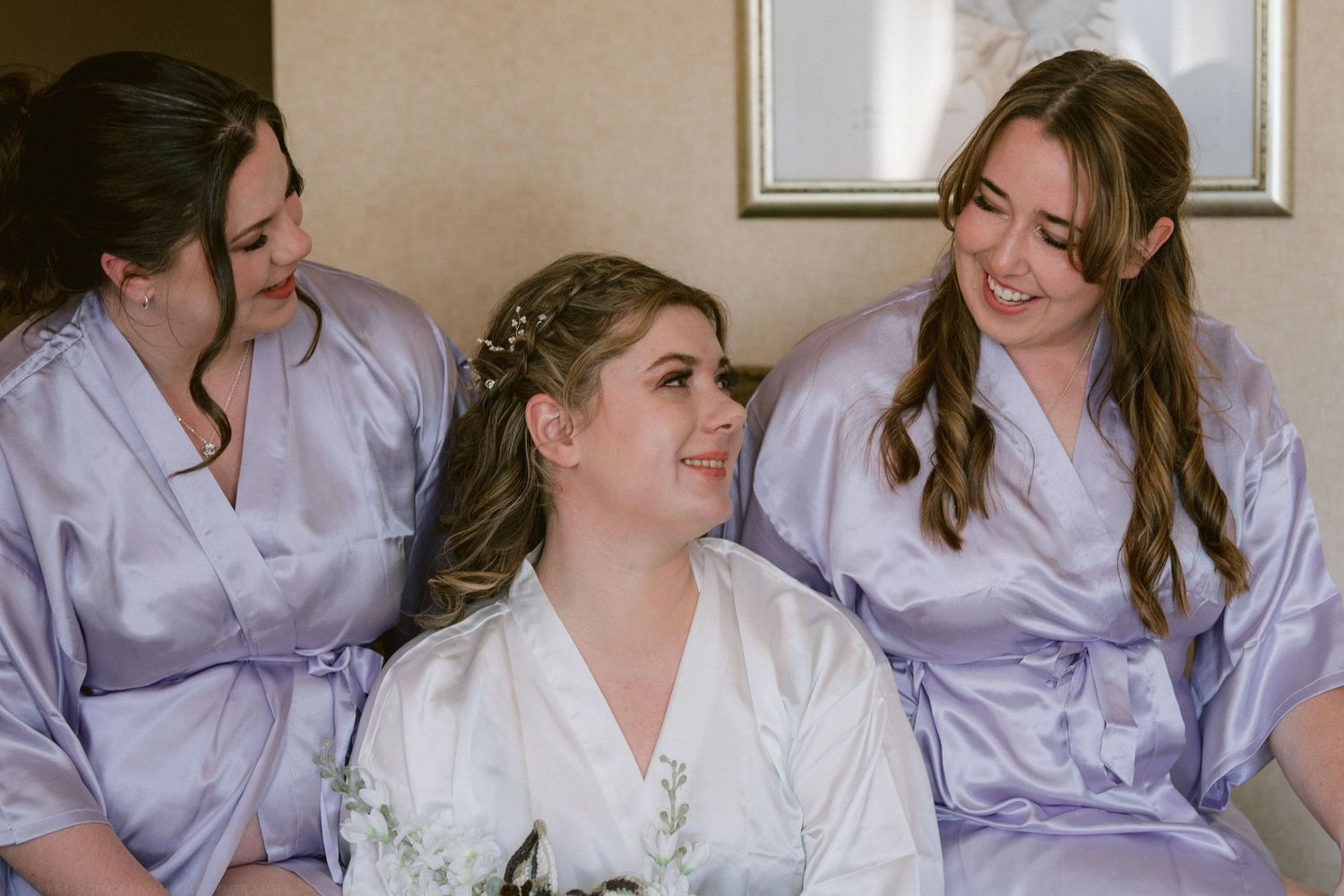 Bride in white robe smiling with two bridesmaids in lavender robes before the wedding.