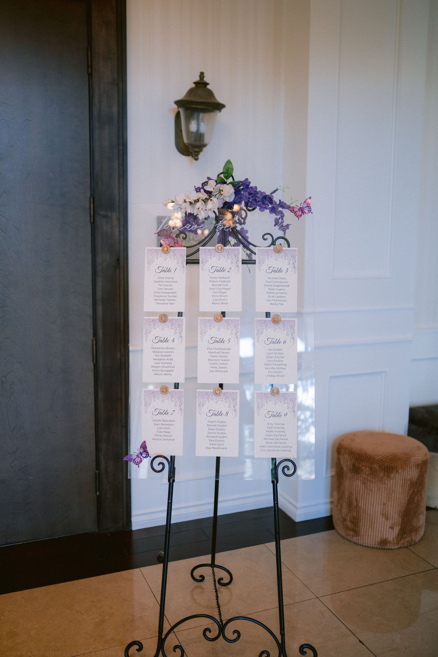 Elegant wedding seating chart display on iron easel decorated with purple and white floral arrangement.