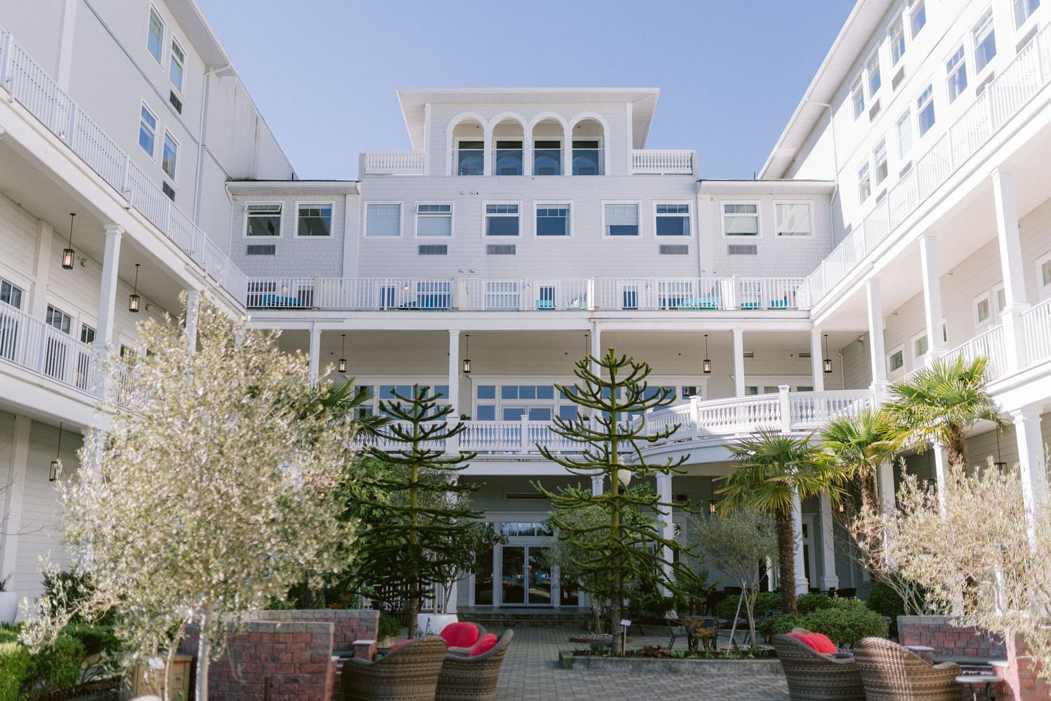 White multi-story hotel with elegant balconies surrounding a lush courtyard with palm trees and potted plants on a sunny day.