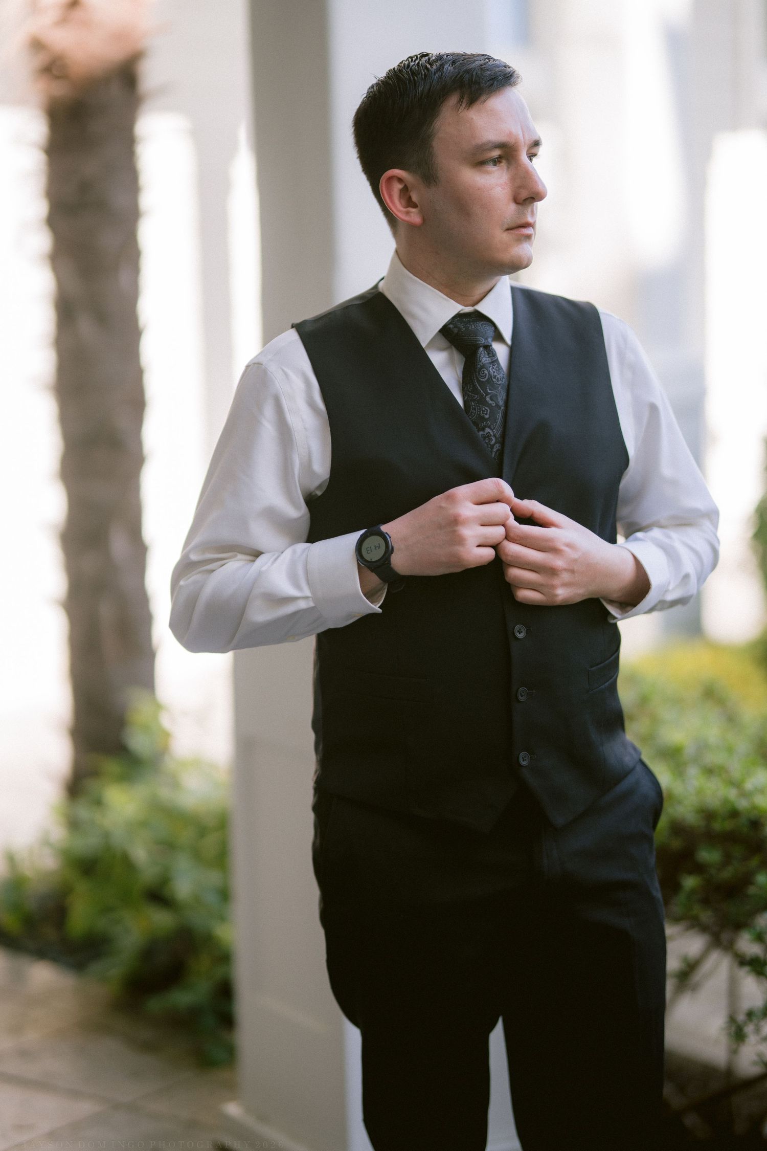 Elegant groom in black vest and tie adjusting his outfit outdoors before wedding ceremony.