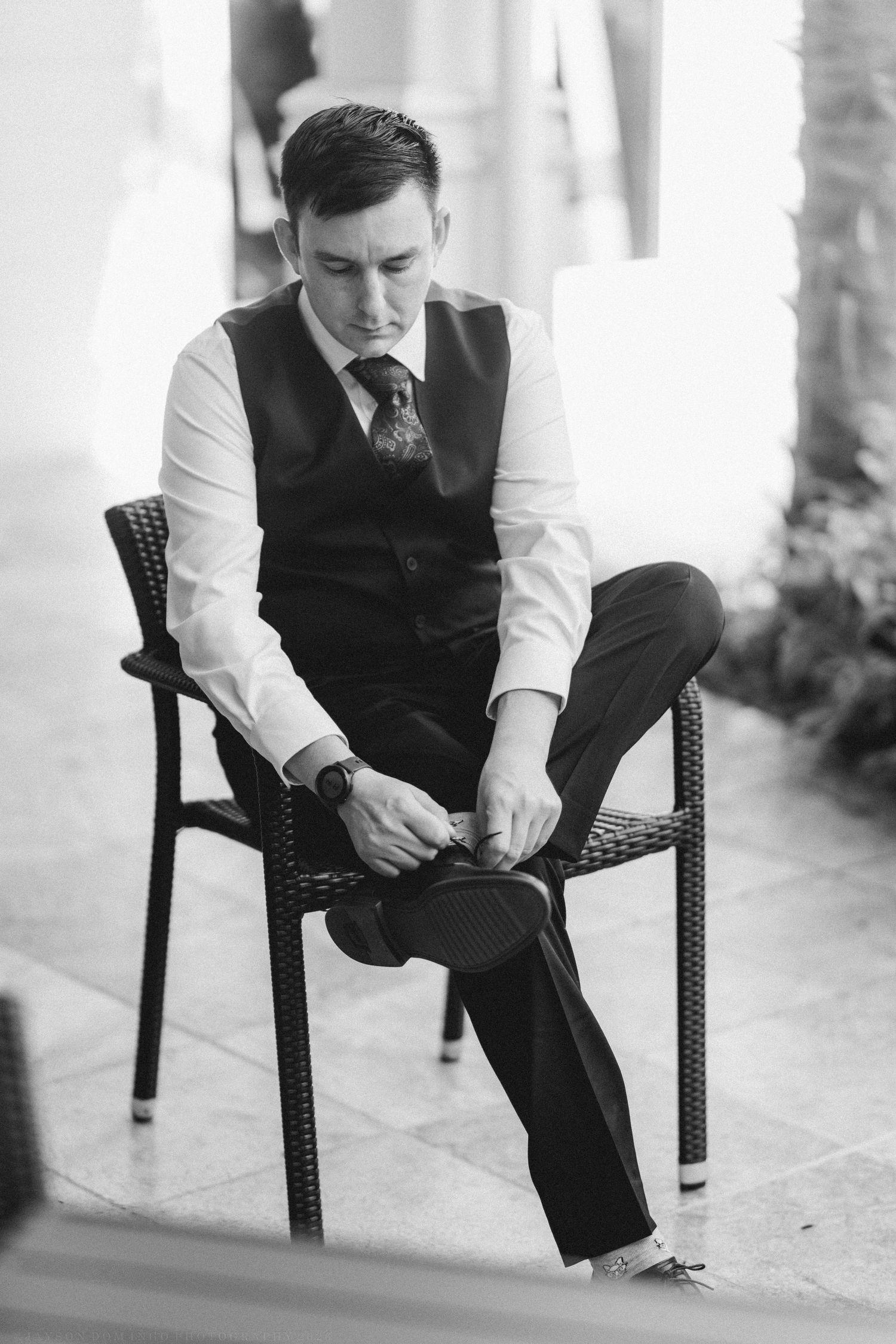 Black and white photo of a well-dressed groom in vest and tie sitting on chair fastening his shoe before wedding ceremony.
