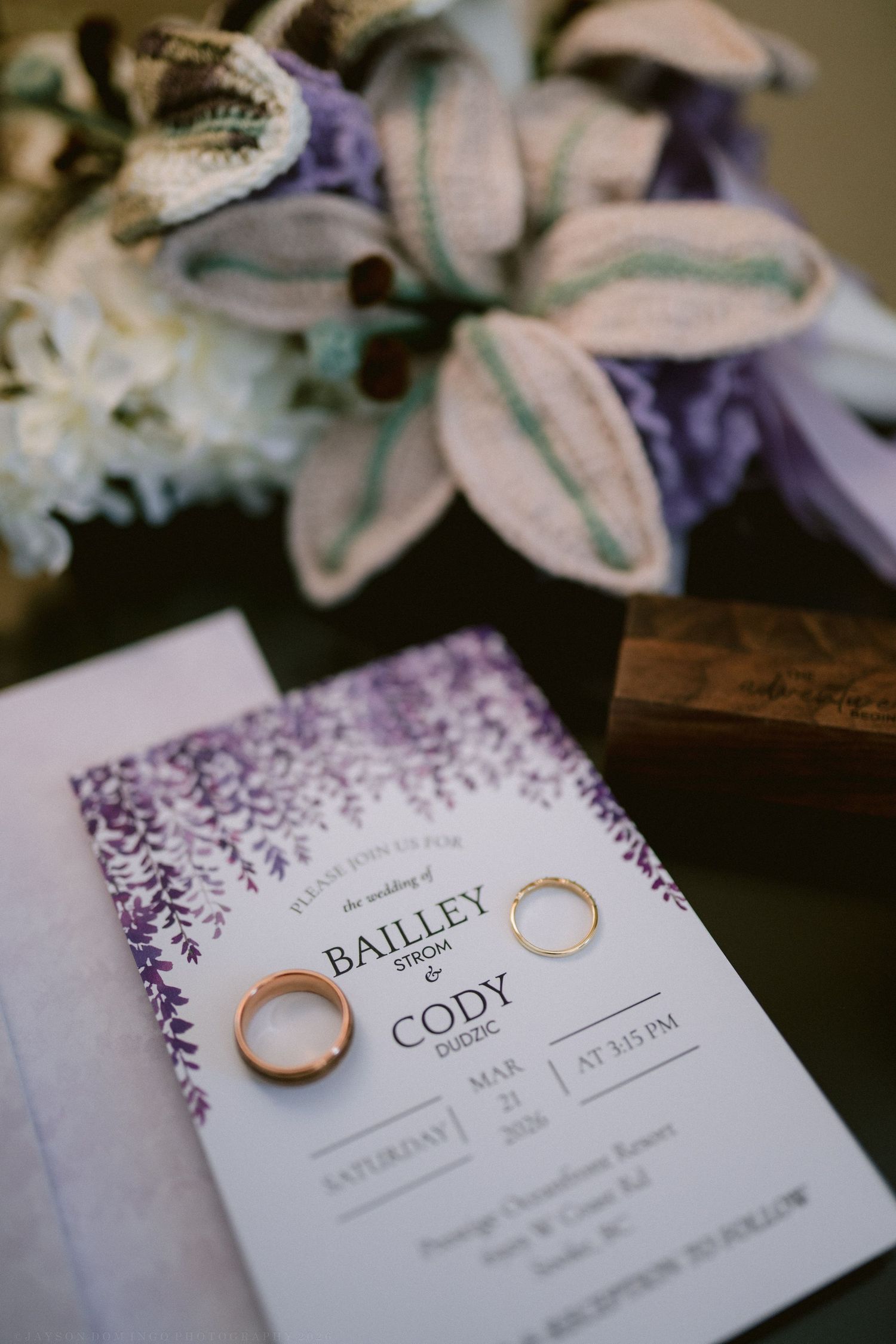 Wedding invitation for Bailley and Cody on a purple floral card with two gold bands and a bouquet nearby.