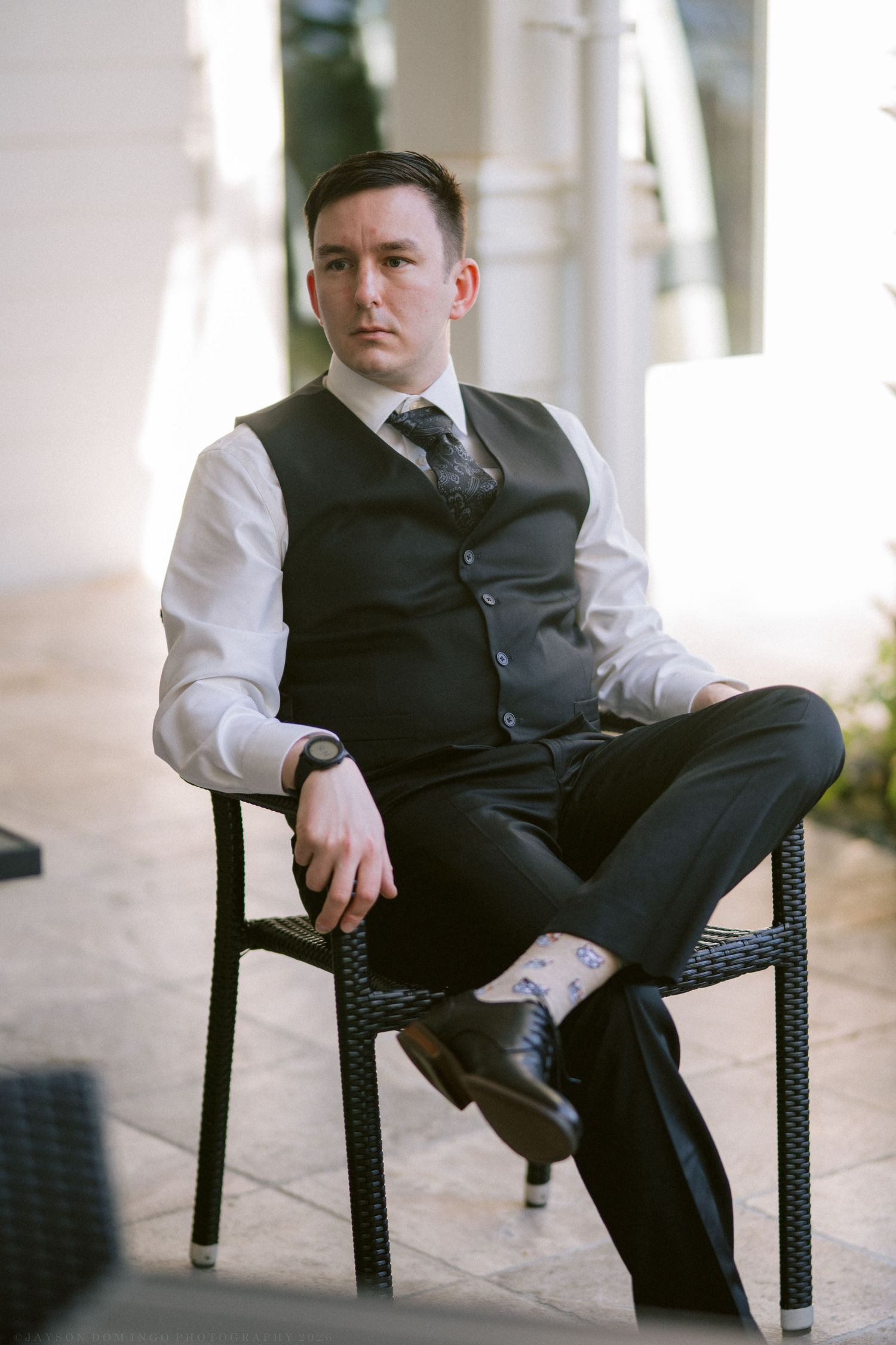 Dapper groom in black suit vest sitting confidently in wicker chair at outdoor wedding venue.