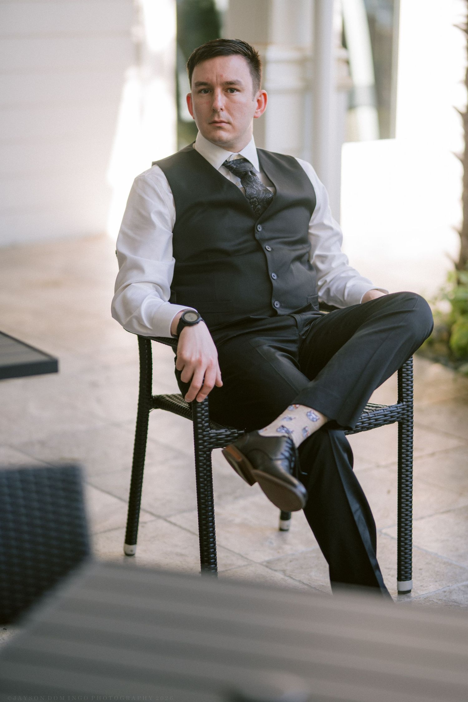 Stylish groom in black waistcoat and tie seated in wicker chair at sunlit wedding venue.