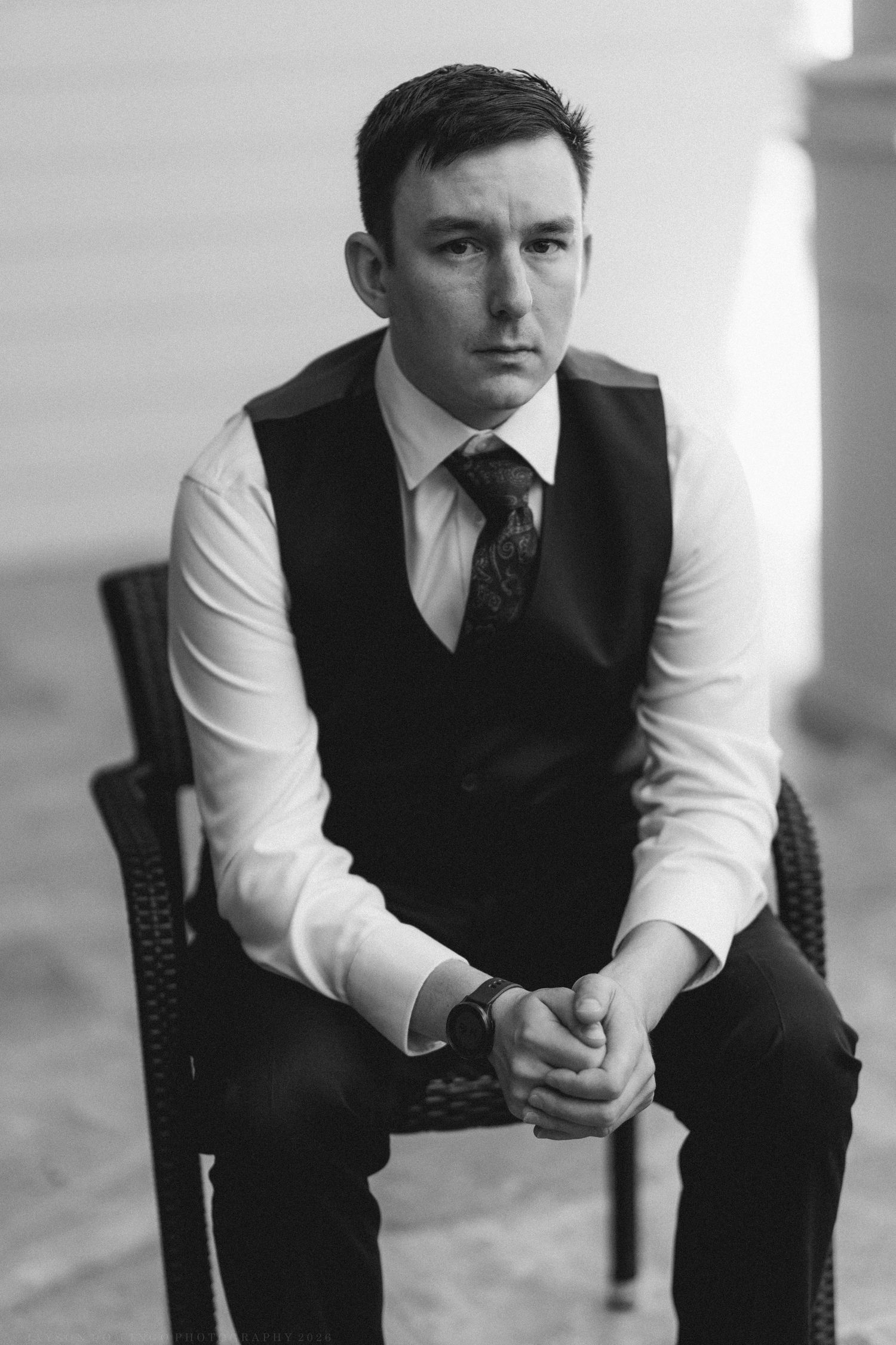 Black and white portrait of a groom in a vest and tie sitting in a chair with hands clasped.