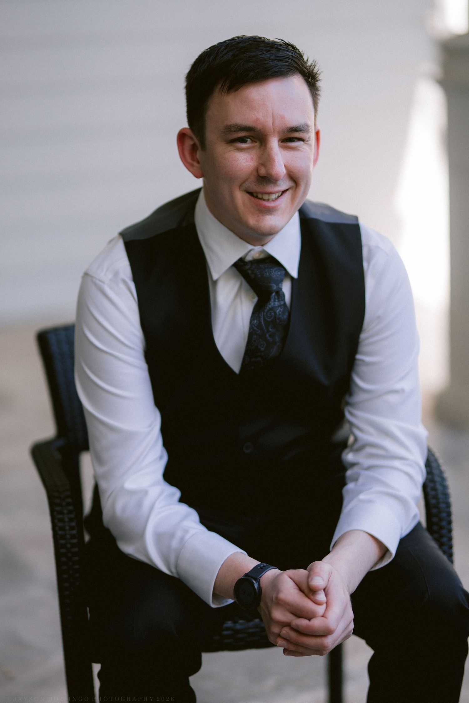 Smiling groom in black vest and tie sitting relaxed in a chair before his wedding ceremony.