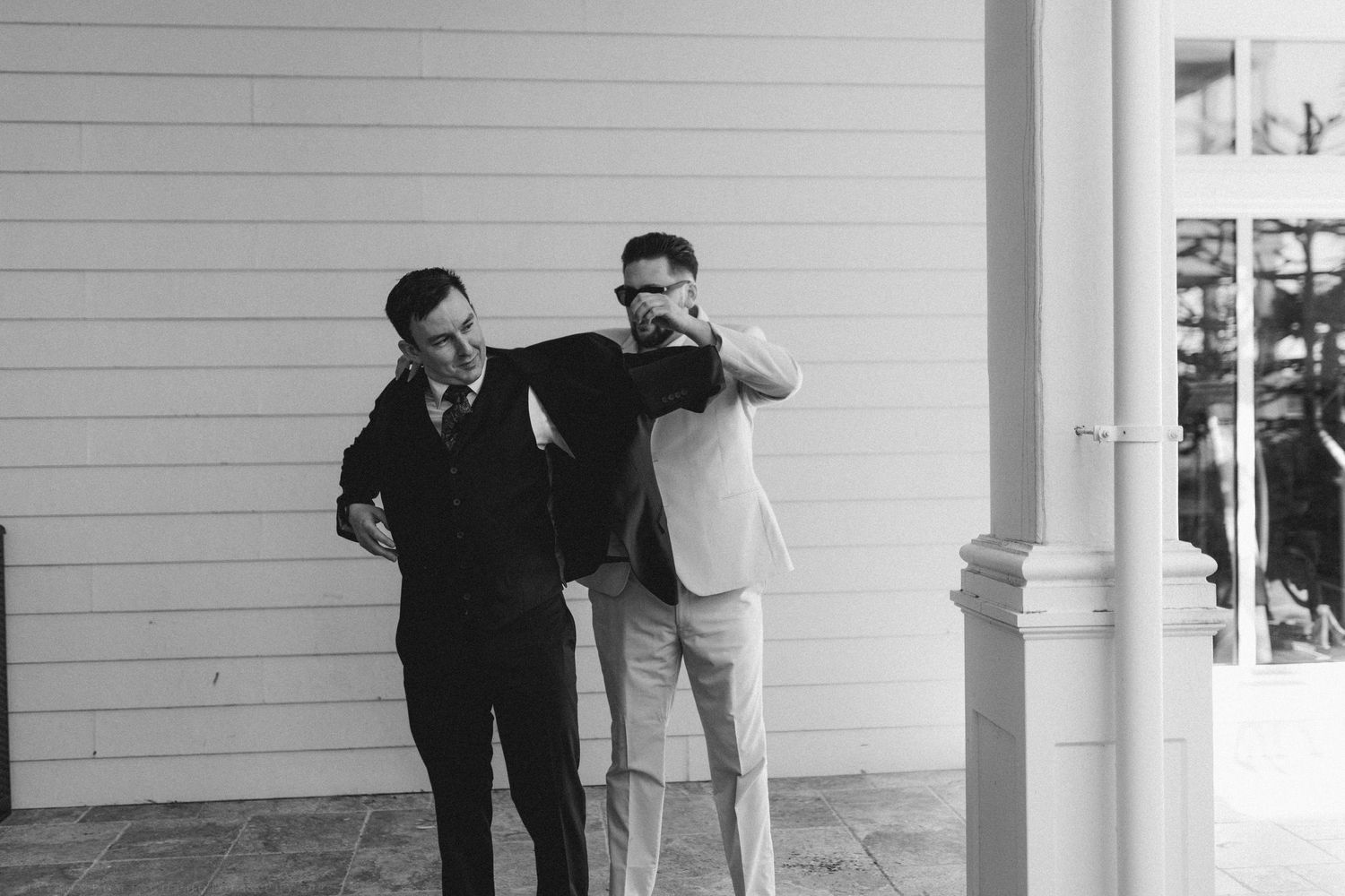 Two men in formal attire laugh together outside a white wooden building in a black and white photo.