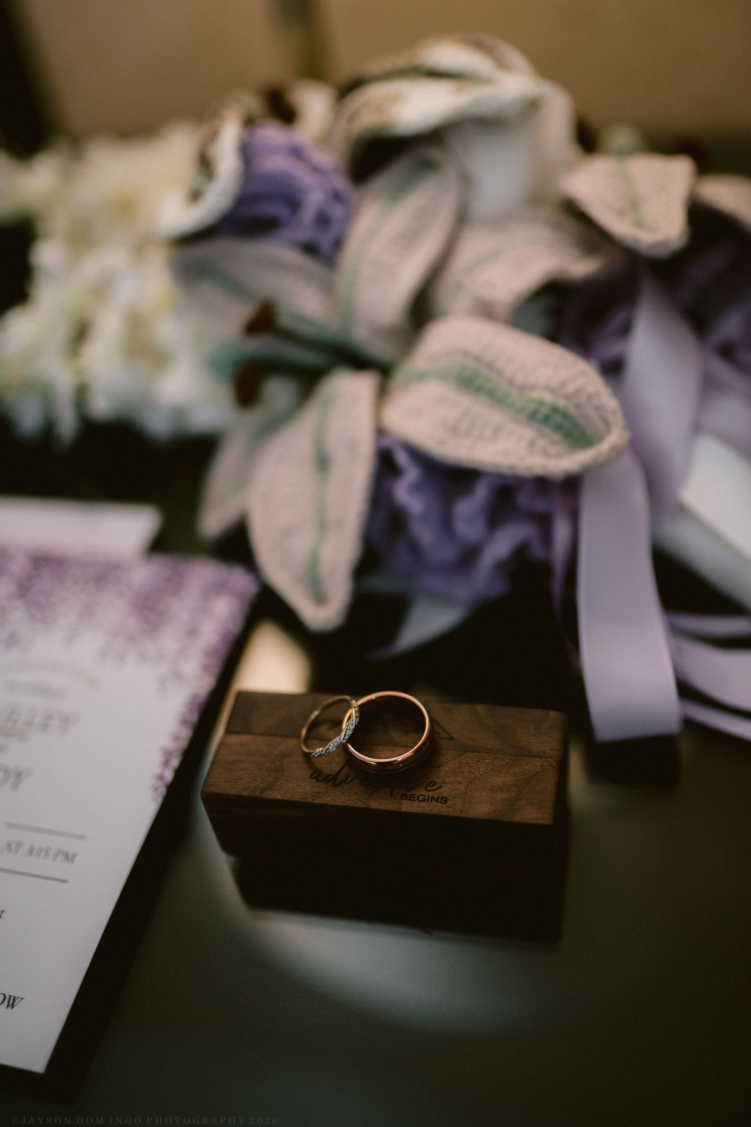 Two gold wedding rings rest on a wooden box beside purple floral bouquet and wedding invitation.