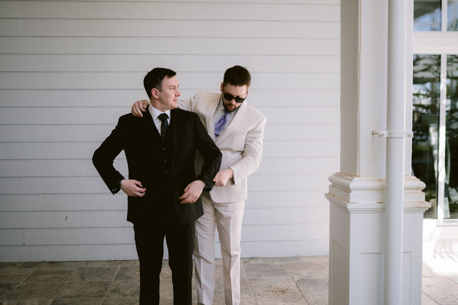 Groom and groomsman sharing a candid moment outside white colonial wedding venue.