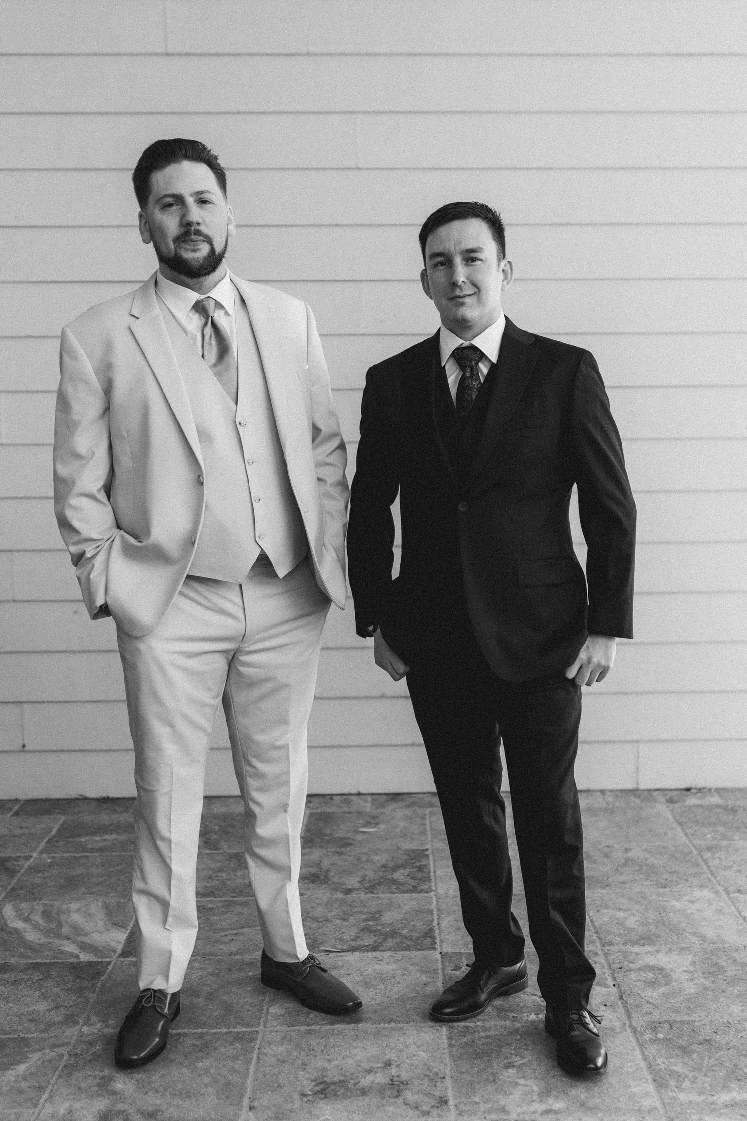 Black and white photo of two groomsmen in suits standing against a white shiplap wall.