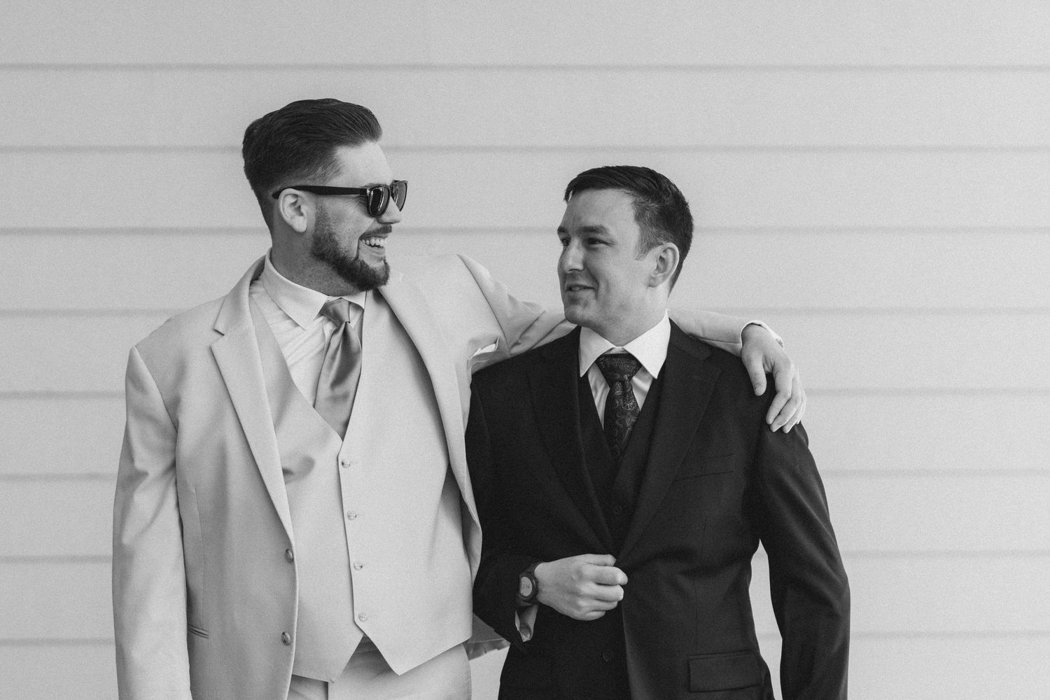 Two men in suits smile and embrace outdoors in a black and white wedding photo.