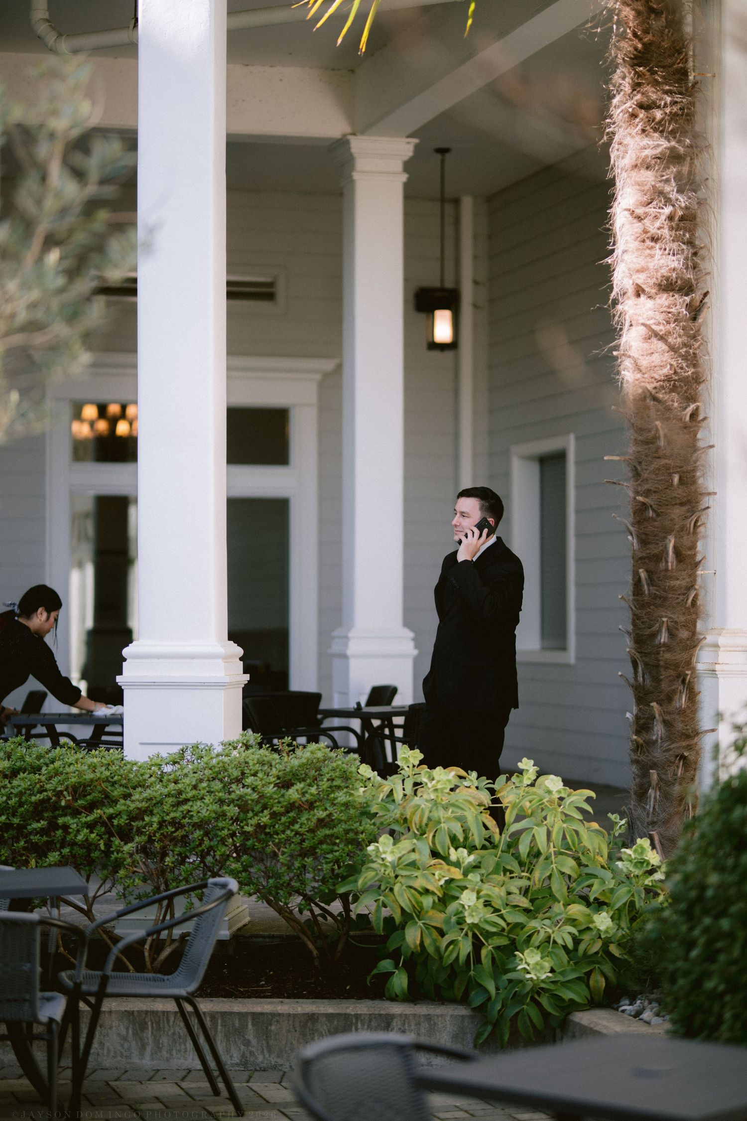 A man in a black suit talks on his phone on an elegant venue porch.