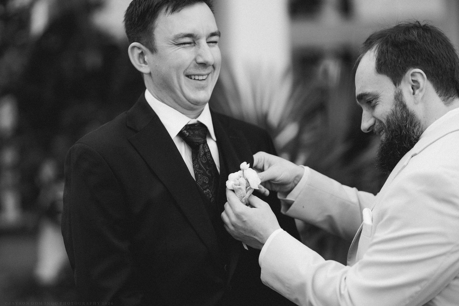 A bearded groomsman pins a boutonniere on a smiling groom in black and white photo.