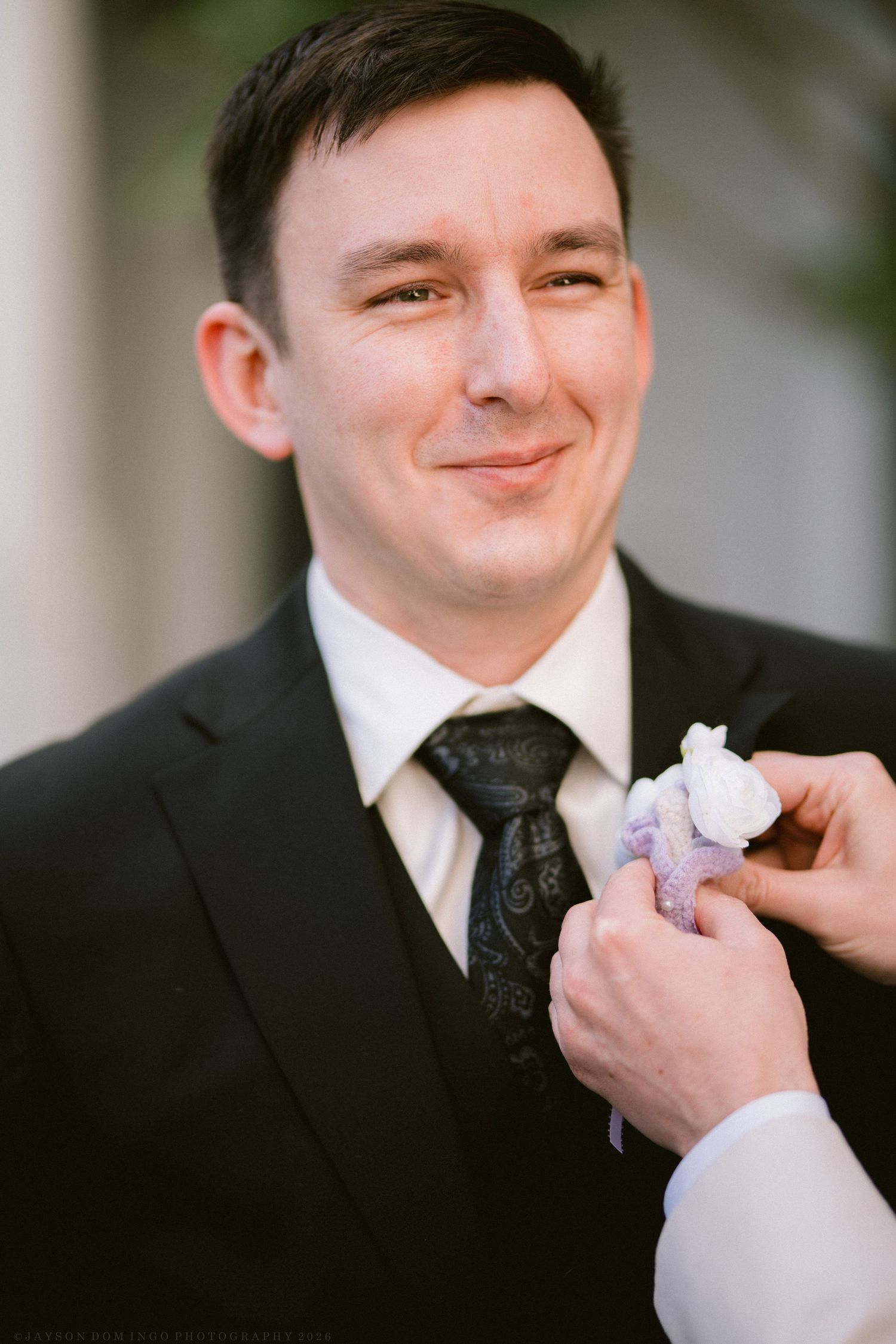 Smiling groom in black suit having white floral boutonniere pinned to his lapel.