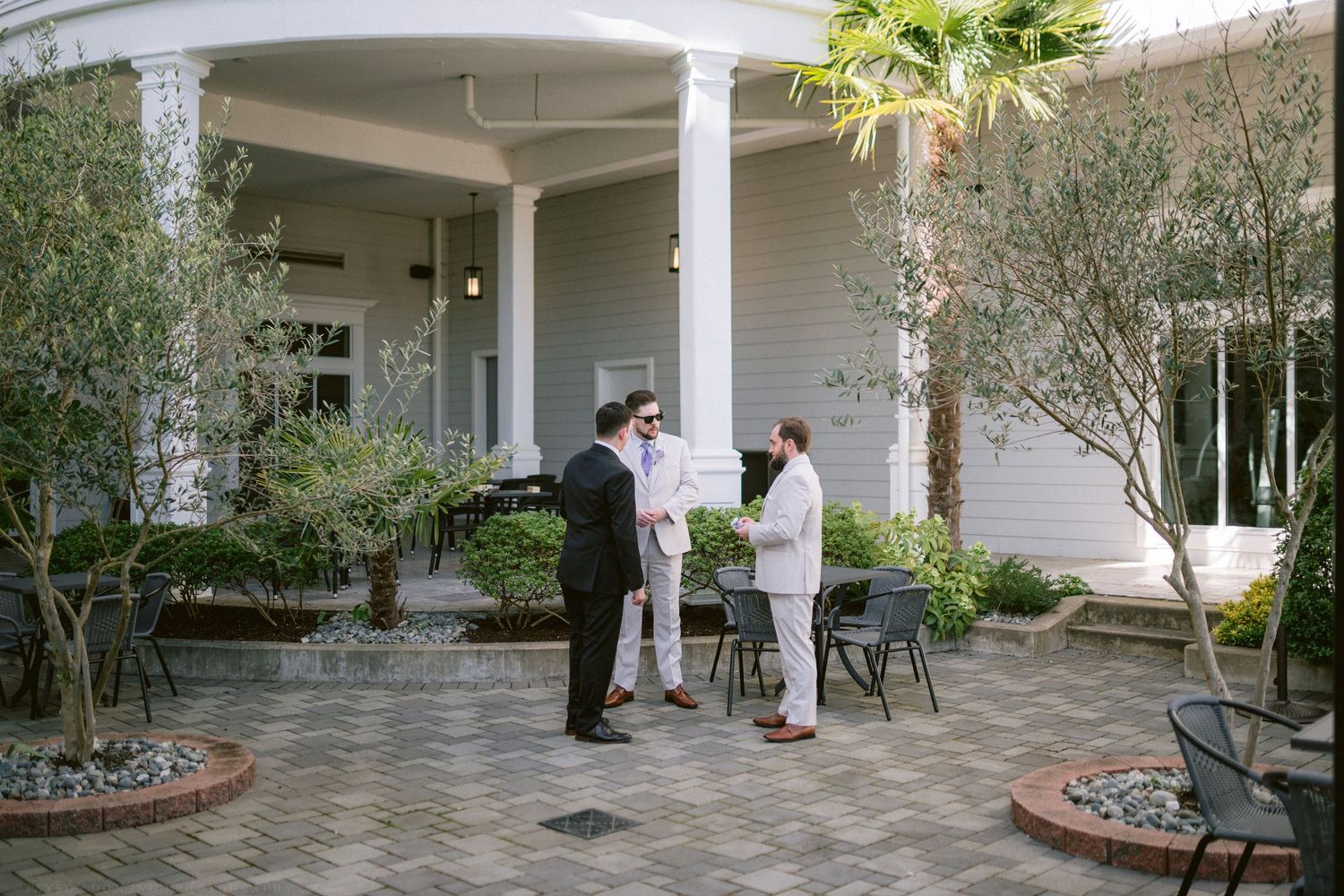 Three men in suits conversing in a courtyard outside an elegant white colonial-style building.