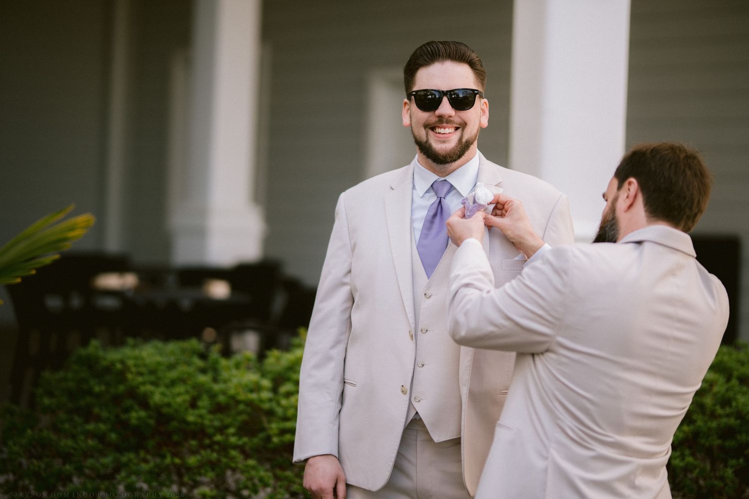 A smiling groom in a tan suit and sunglasses gets his tie adjusted outdoors.
