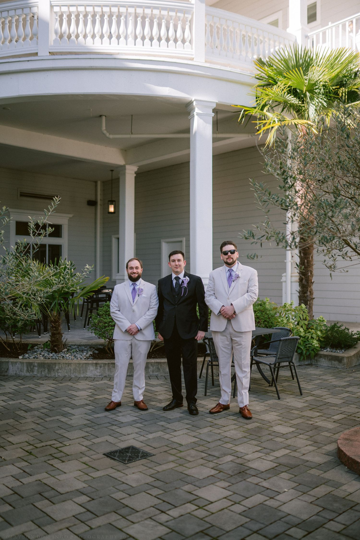 Groom in black suit flanked by two groomsmen in light gray suits at elegant wedding venue.