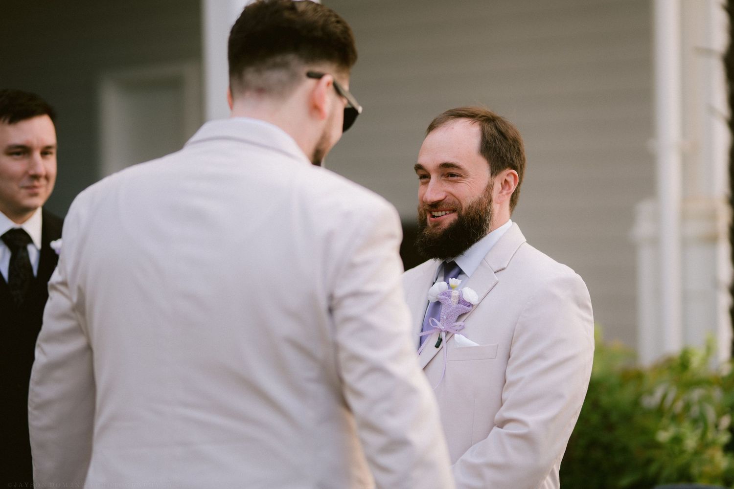 Groom smiles warmly at partner during outdoor wedding ceremony wearing beige suit with floral boutonniere.