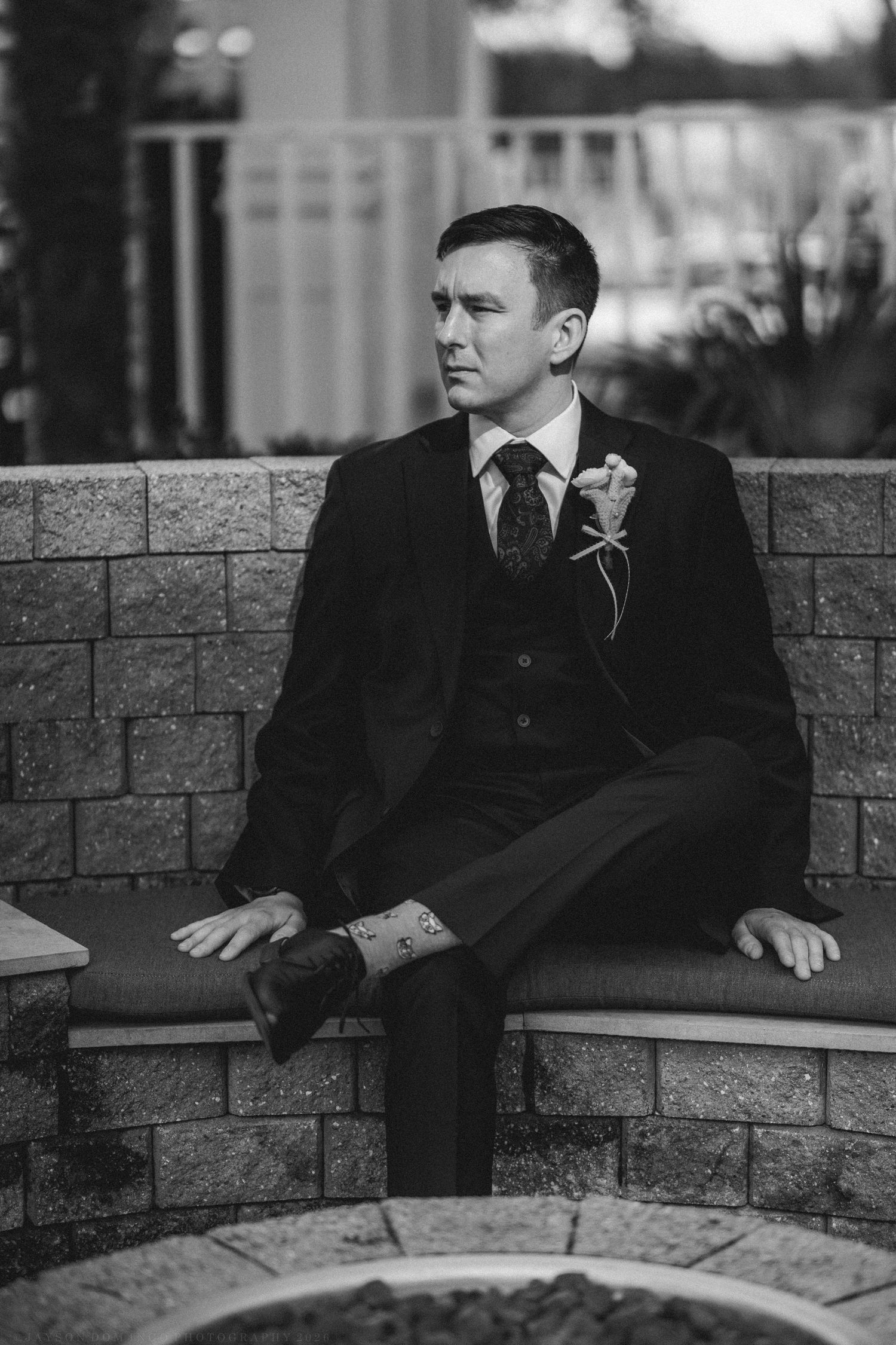 Dapper groom in black suit with boutonniere sitting on stone bench in black and white wedding portrait.