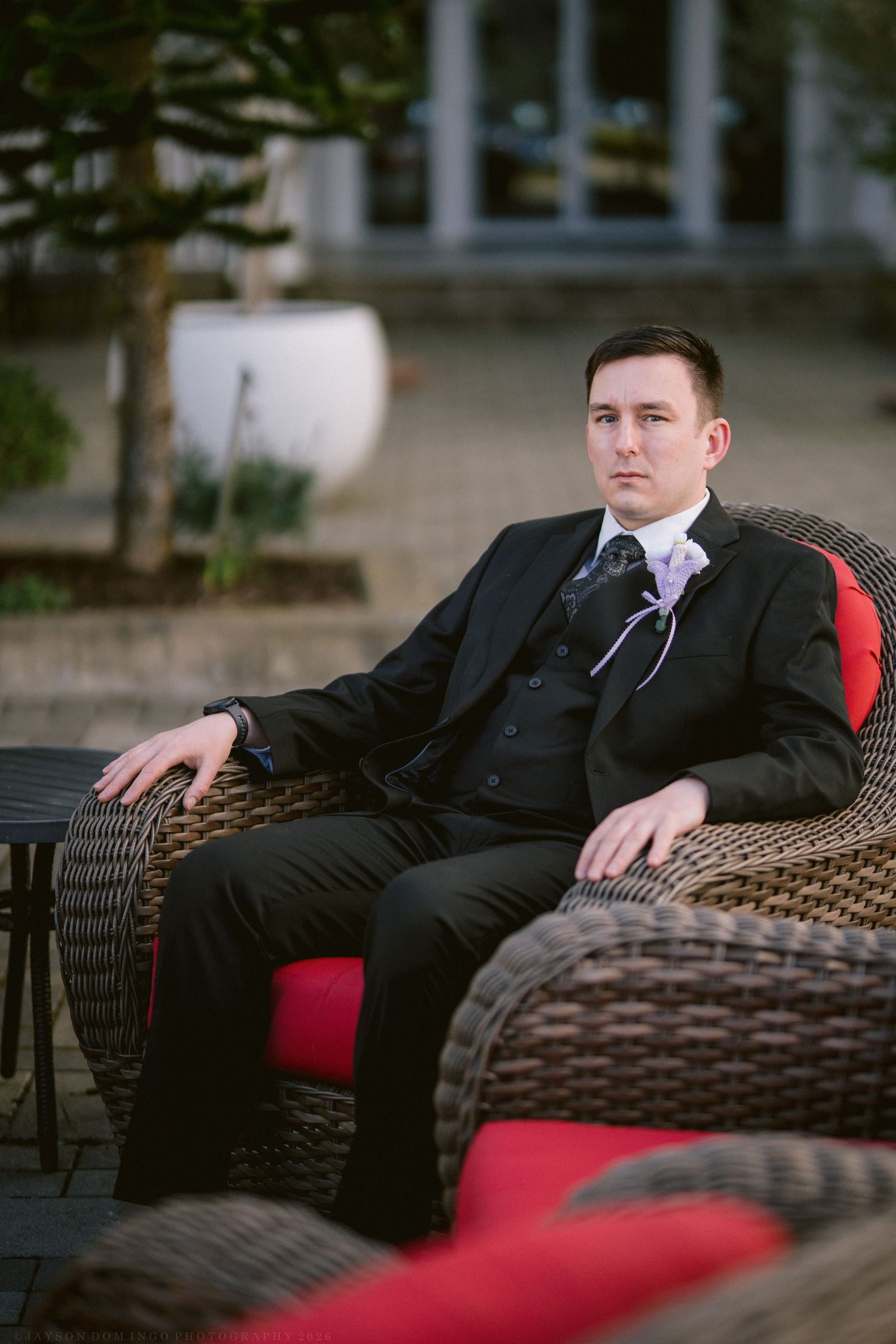 Groom in a black suit relaxing in a wicker chair with red cushions at an outdoor venue.
