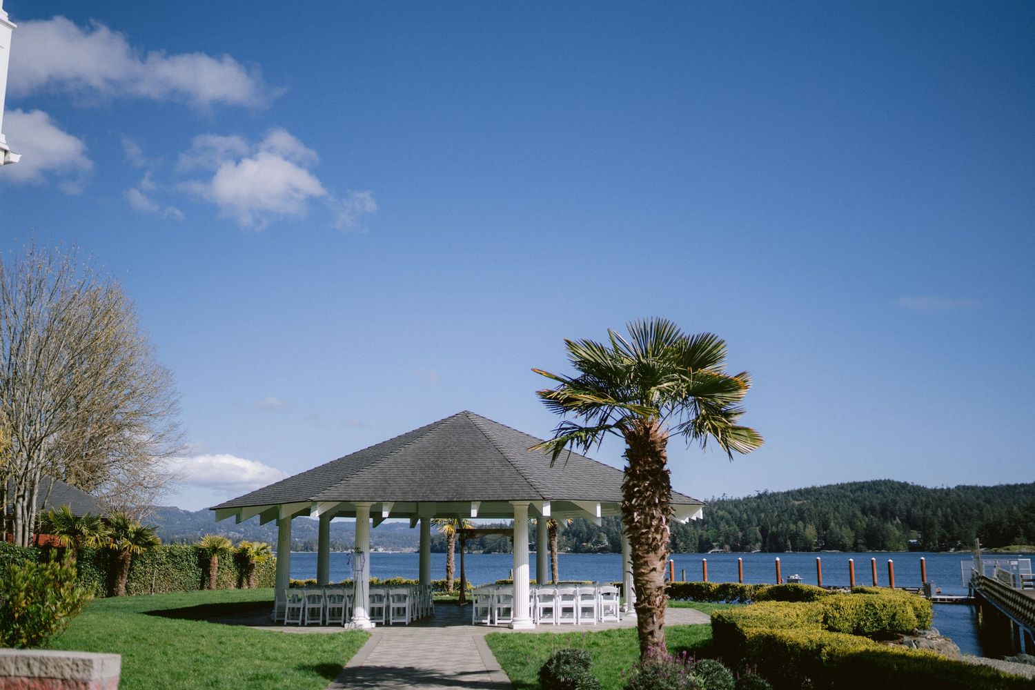 Elegant waterfront gazebo with palm tree and mountain views under blue skies, perfect for outdoor wedding ceremonies.