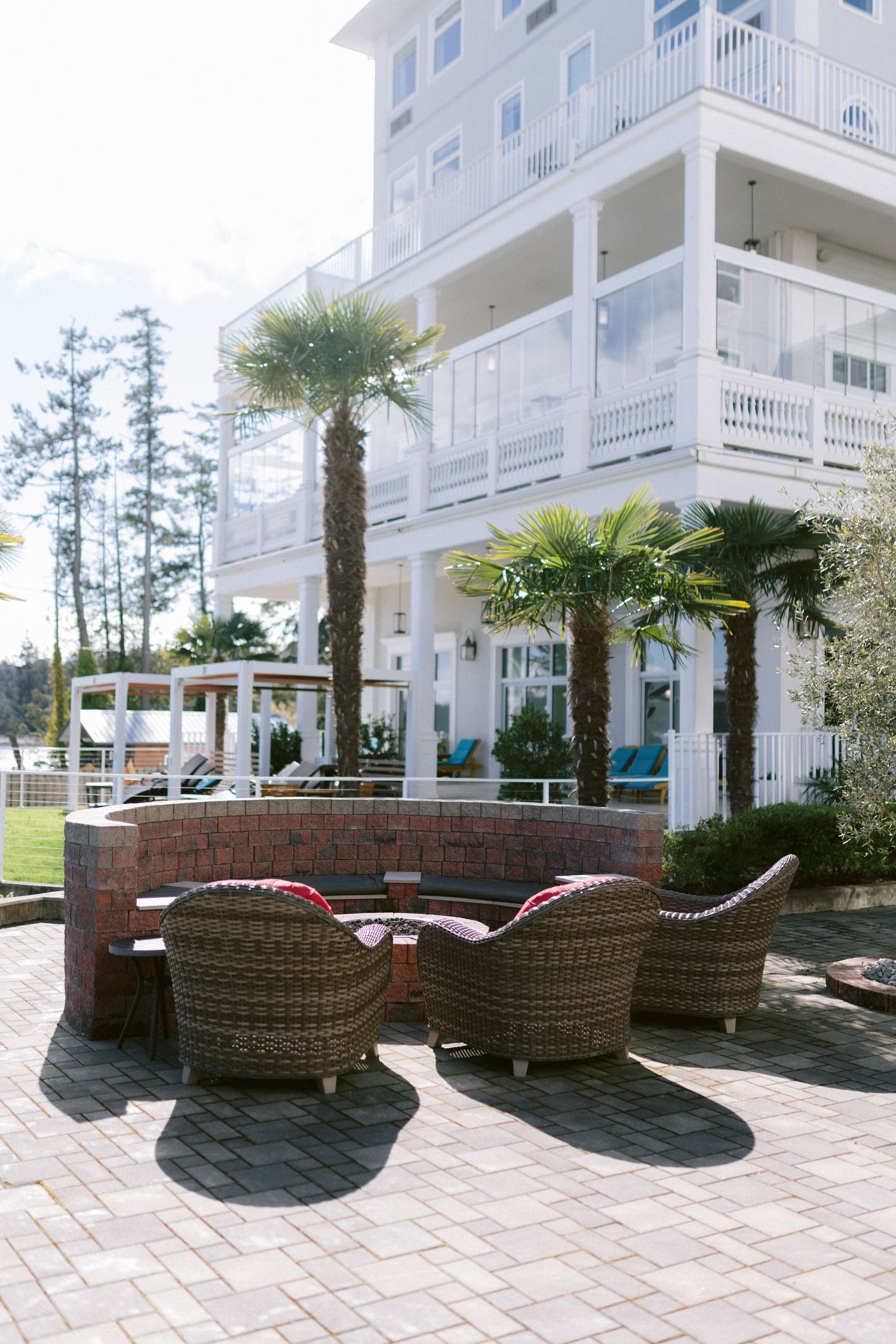 Wicker chairs on brick patio outside white multi-story hotel with palm trees and balconies on sunny day.
