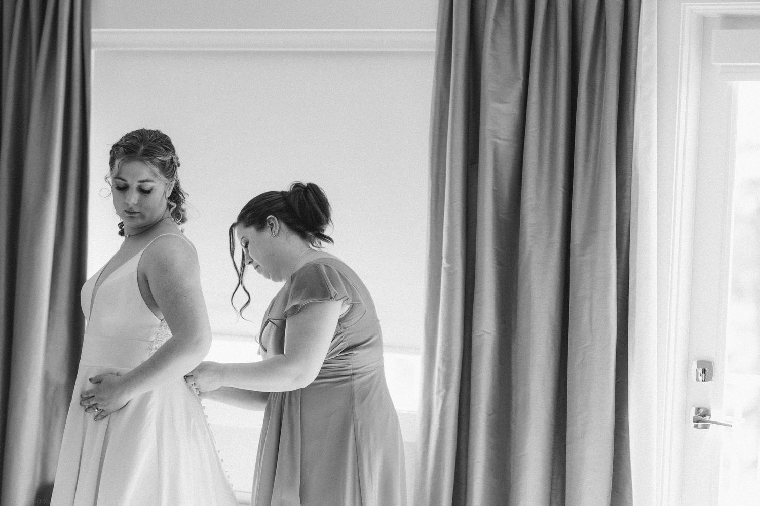 Black and white photo of a bridesmaid helping the bride with her wedding dress near a window.