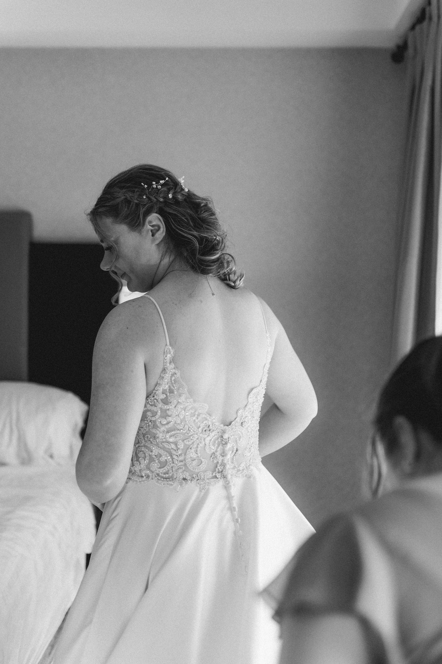 Bride in lace wedding gown stands in black and white hotel room during bridal preparations.