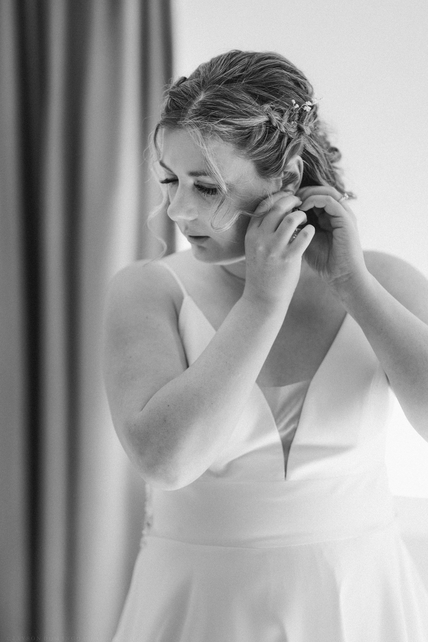 Bride in white wedding gown putting on earrings while getting ready, black and white photo.