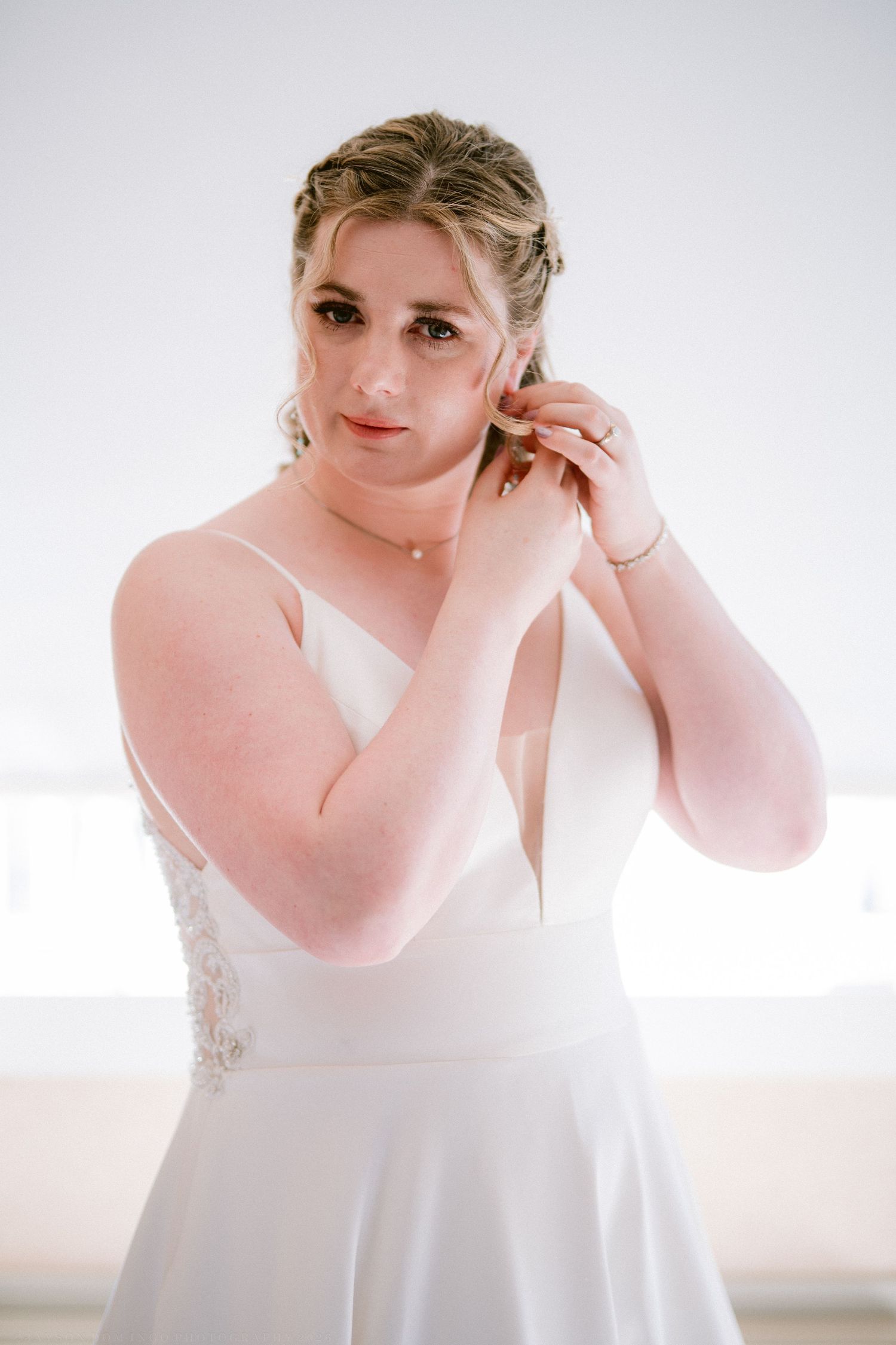 Bride in white gown adjusting earring, lace detail dress, elegant updo hairstyle, soft natural lighting.