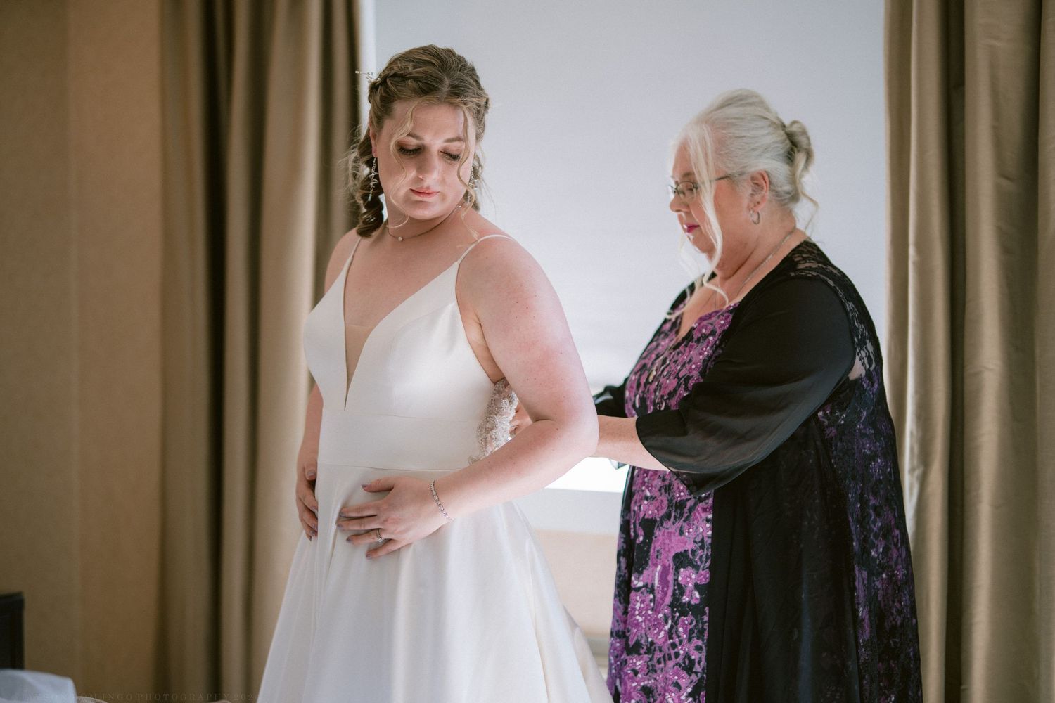 Bride in a white gown being helped into her dress by an older woman in a hotel room.