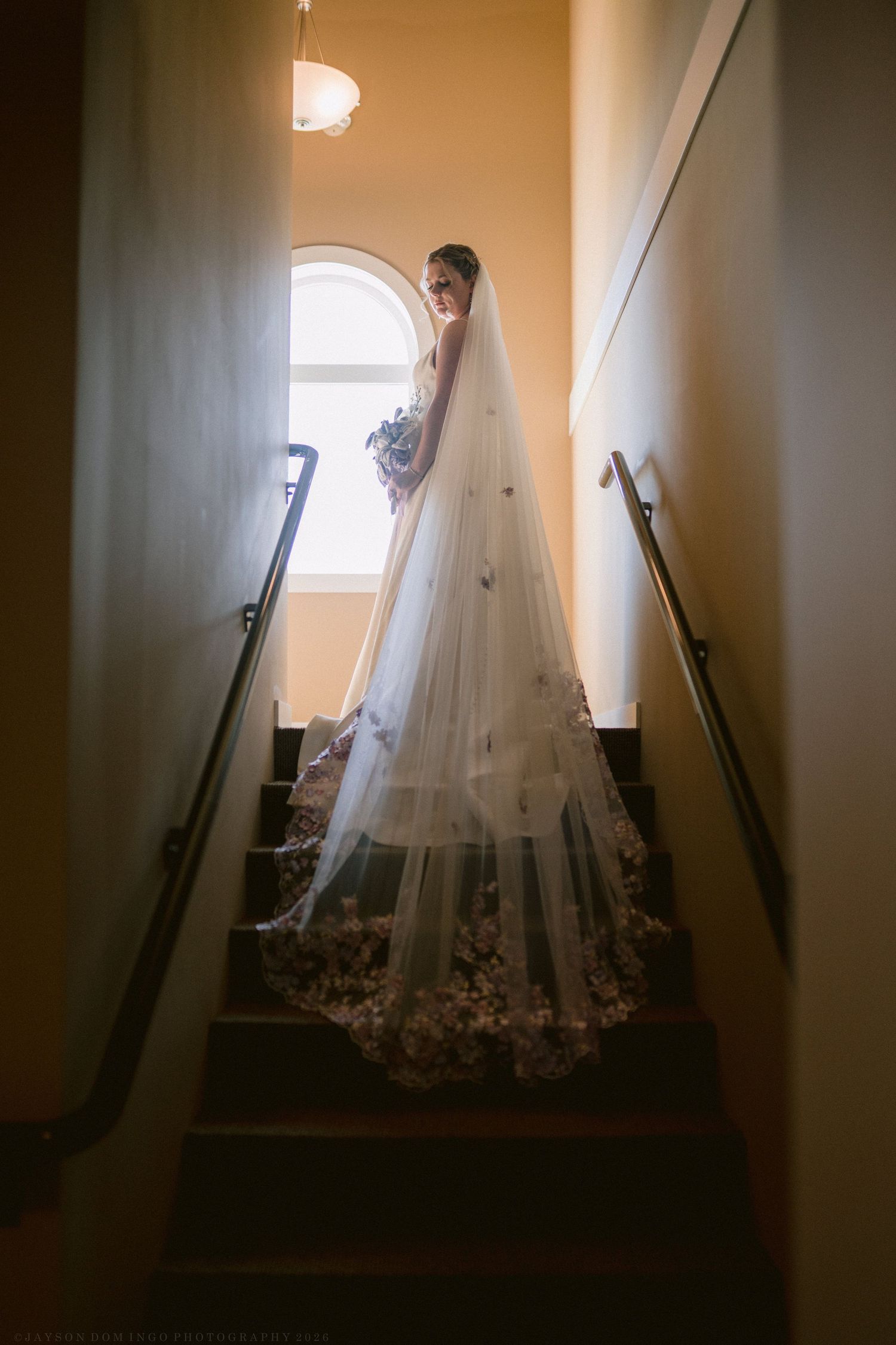 Bride in flowing gown with floral cathedral veil standing gracefully on a staircase by arched window.