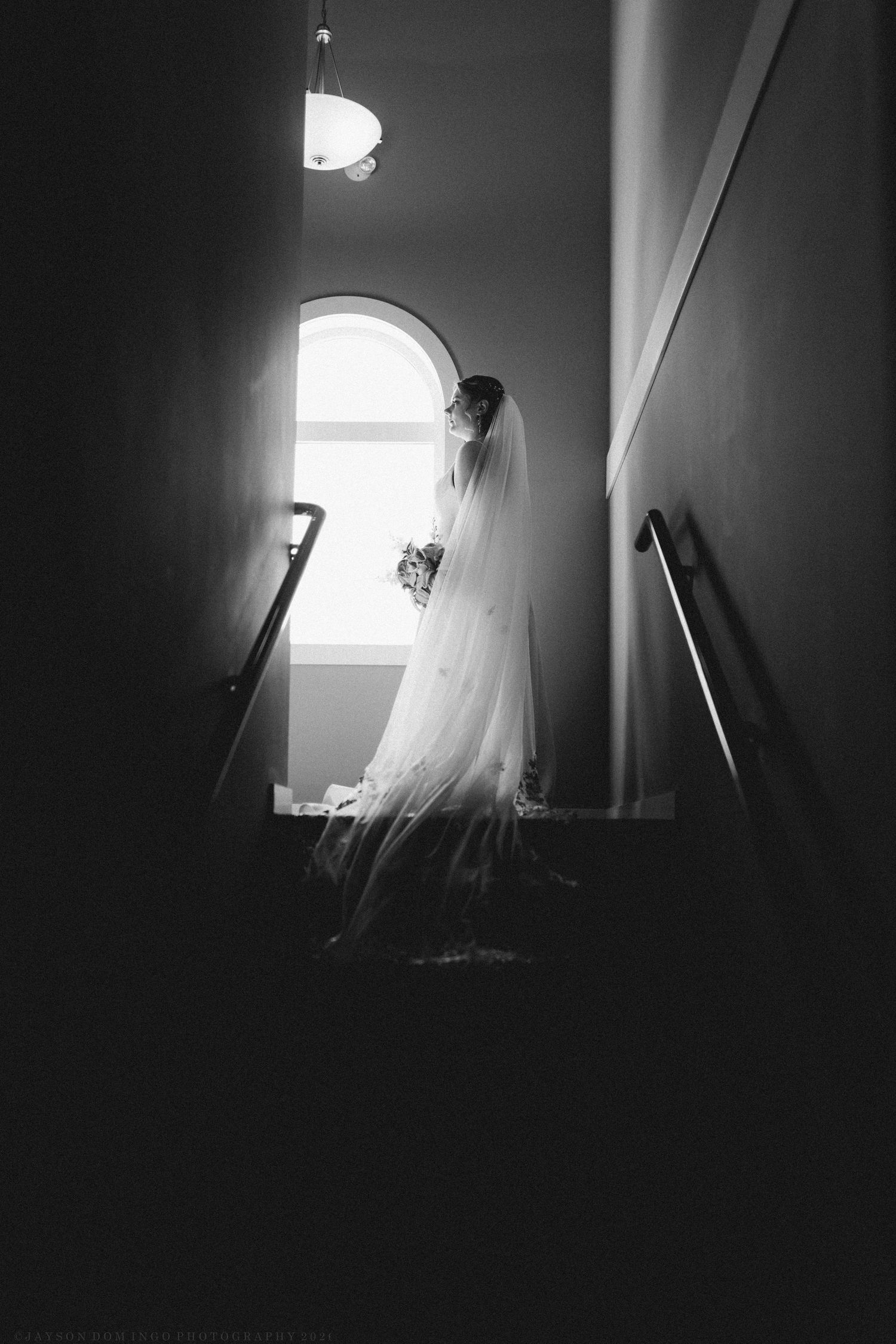 Black and white silhouette of bride in wedding gown and veil standing by arched staircase window.