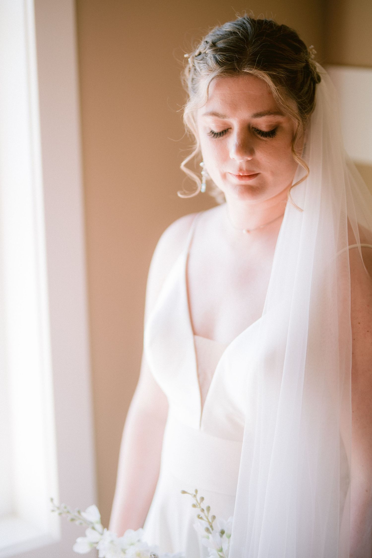Bride in white gown and veil gazes down softly near a sunlit window, with delicate flowers in the foreground.