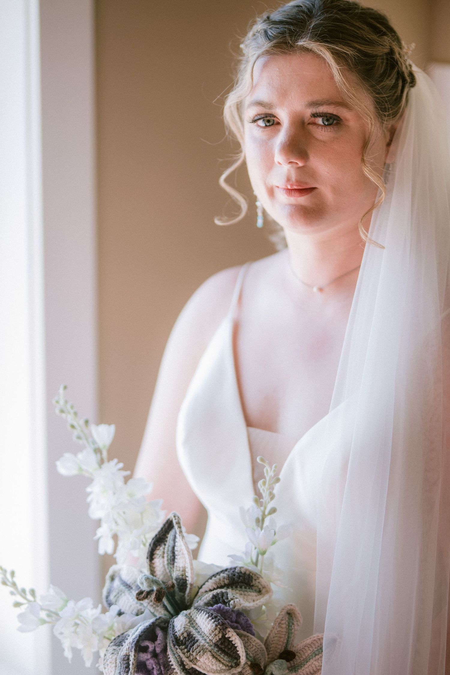 Elegant bride in white gown and veil holding a floral bouquet, standing near a window in soft natural light.
