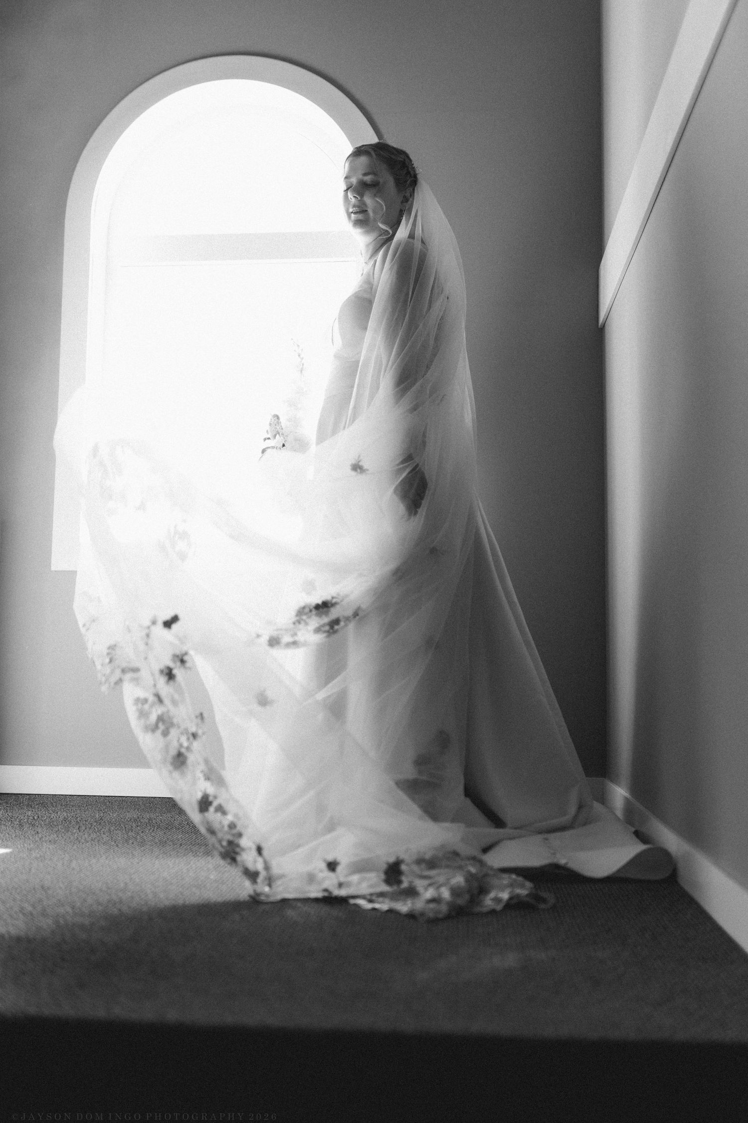 Black and white silhouette of bride in long floral veil standing by arched window in dramatic light.