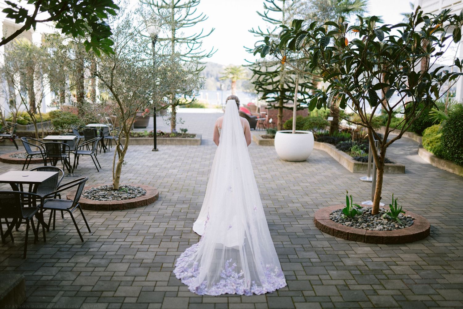 Bride in white wedding gown with long lace veil standing in courtyard surrounded by potted trees.