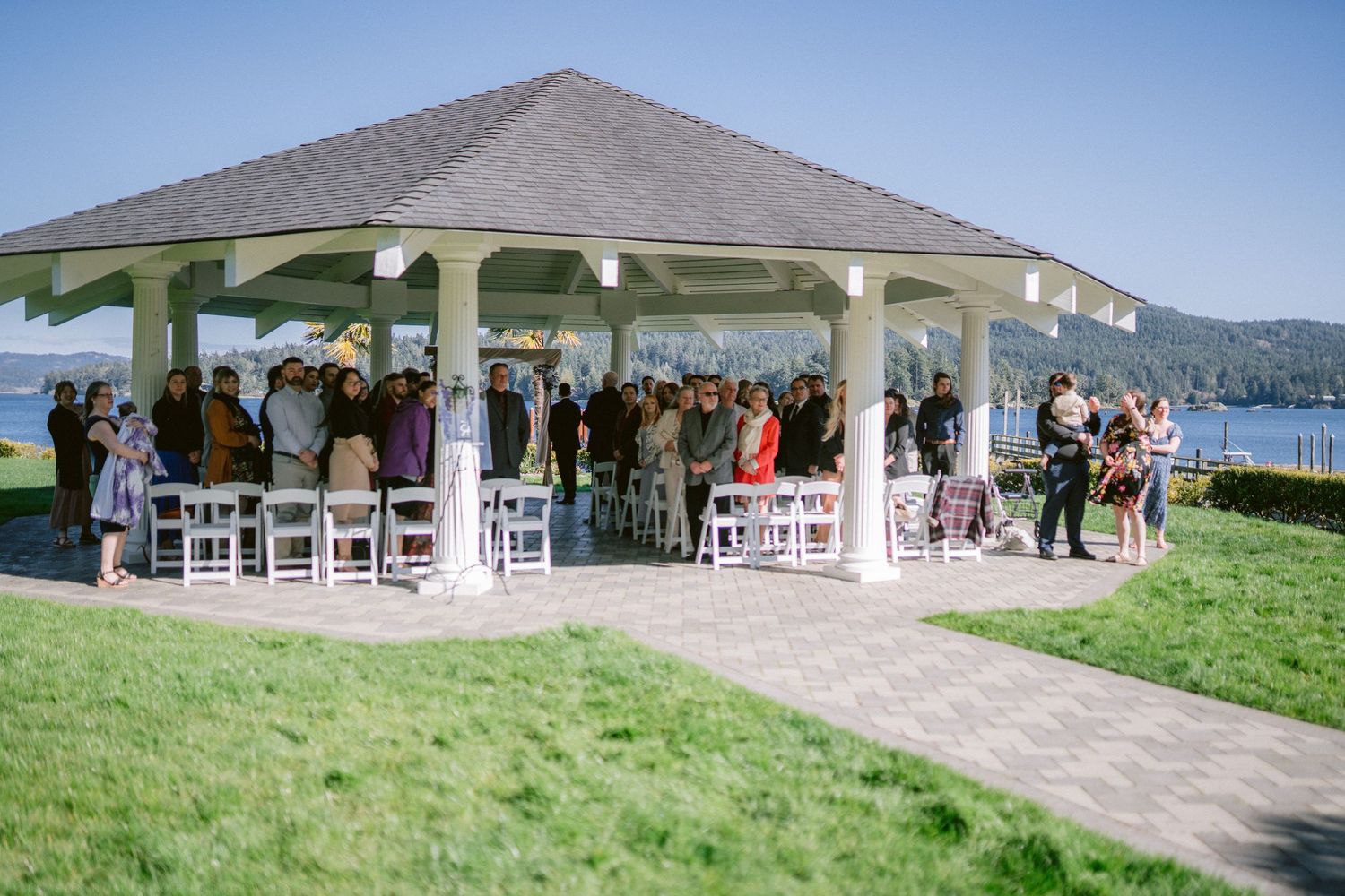 Outdoor wedding ceremony under a gazebo pavilion overlooking a scenic waterfront on a sunny day.