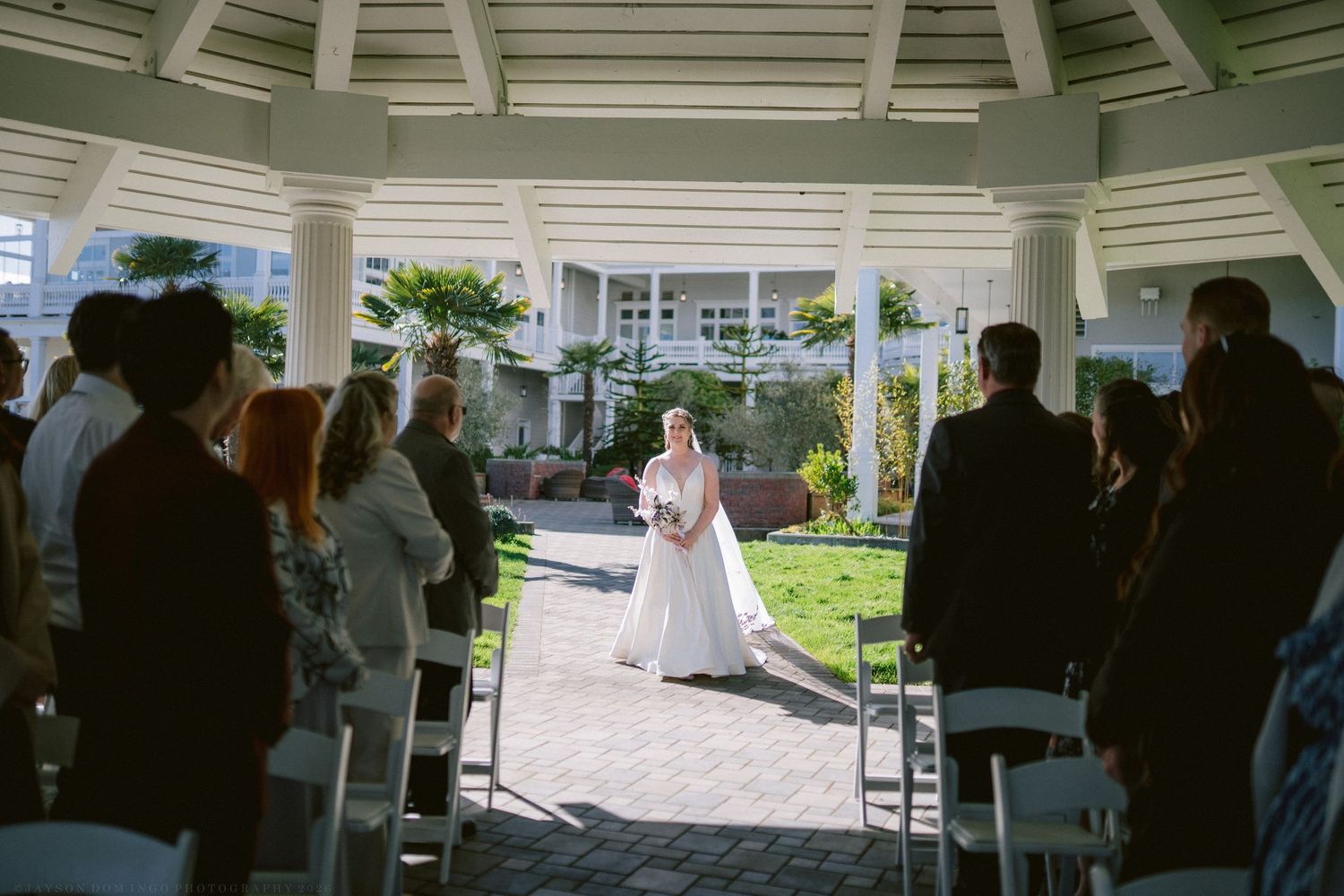 Bride walks down aisle toward gazebo altar at outdoor waterfront wedding ceremony with seated guests.