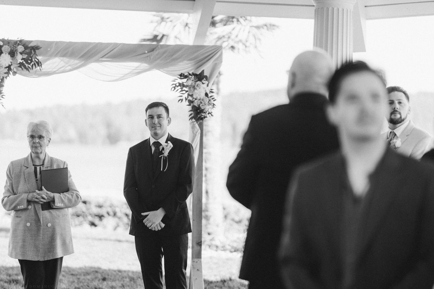 Black and white photo of groom waiting at altar watching bride walk down the aisle outdoors.