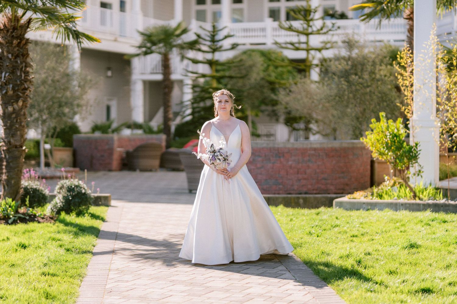 Bride in full ballgown wedding dress holds bouquet while posing in sunny garden of venue.