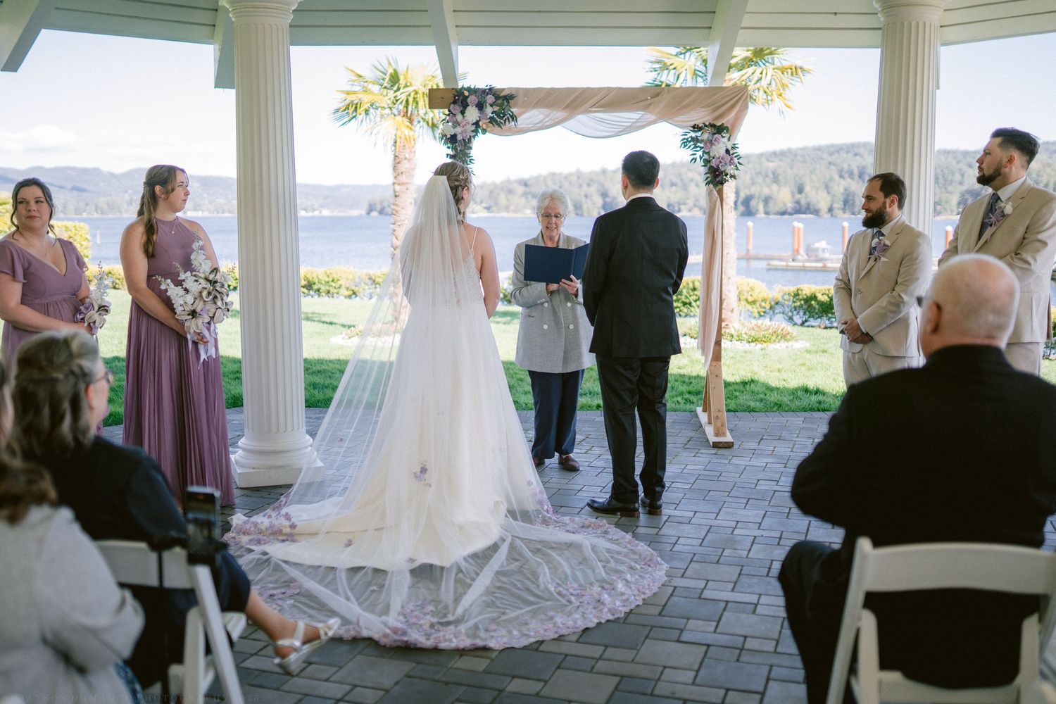 Bride and groom exchange vows under floral arch in gazebo with waterfront view and wedding party.