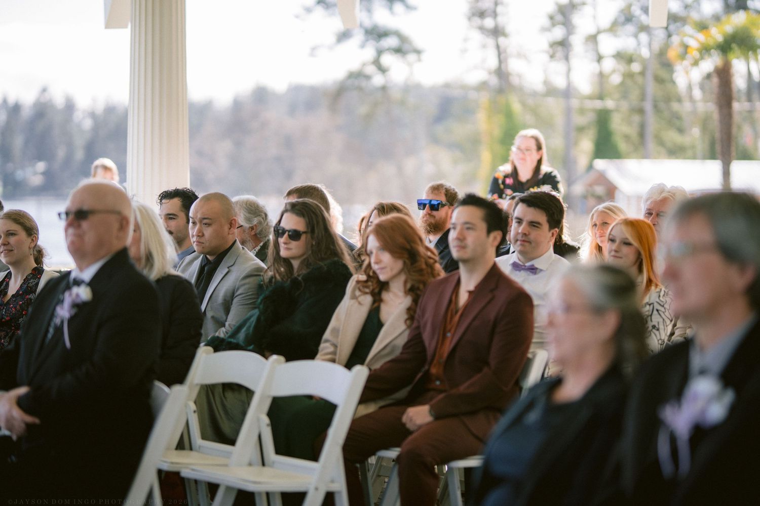 Guests seated in white chairs at an outdoor wedding ceremony, dressed in elegant attire, with trees visible in the background.