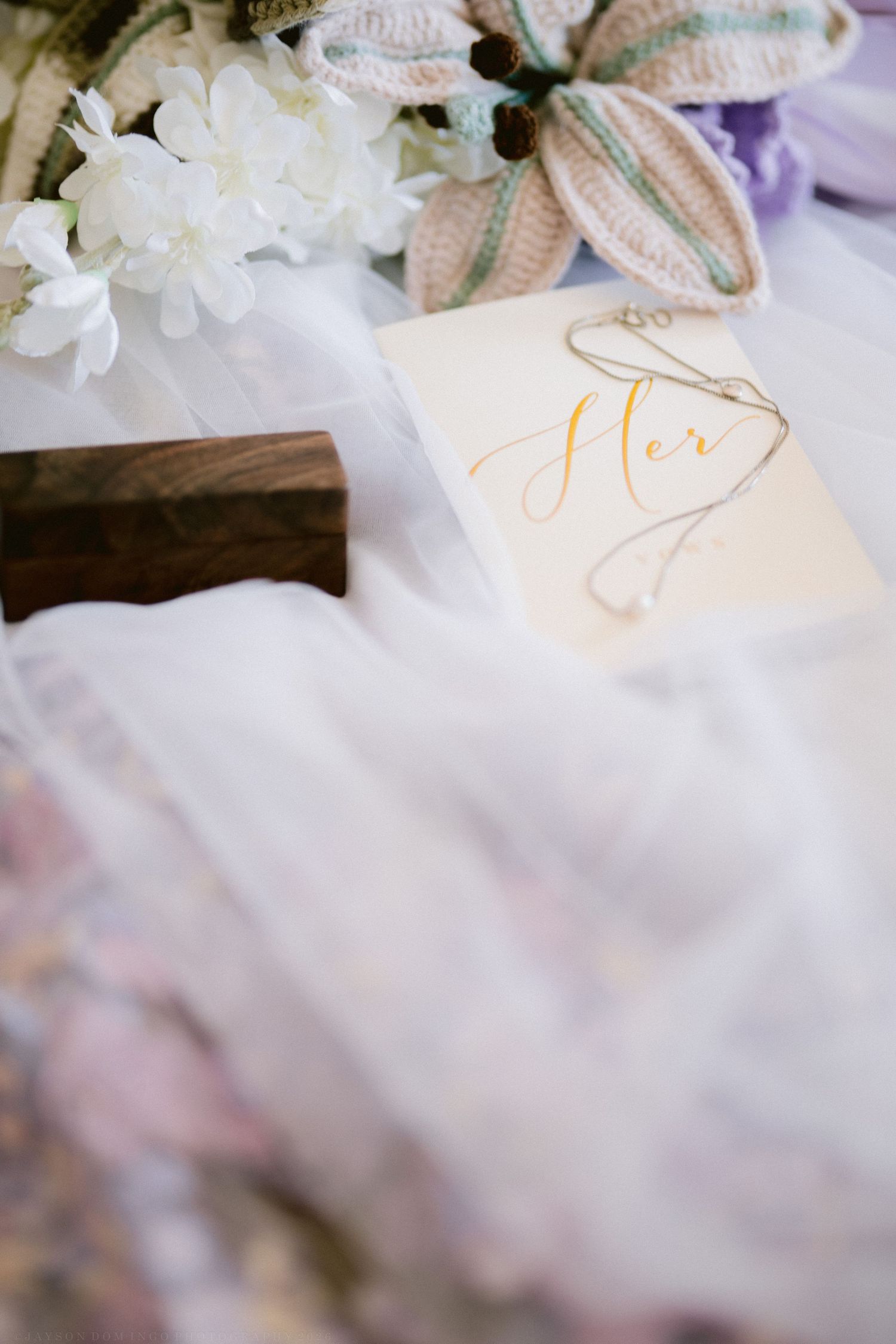 Bridal flat lay with a gold script Her card, white flowers, wooden ring box, and flowing white veil.