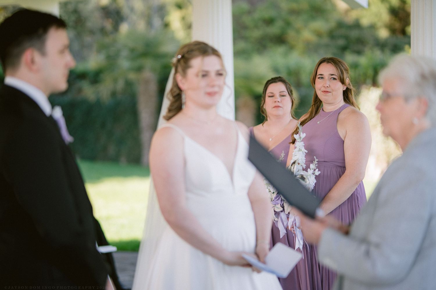 Bride smiling at groom during outdoor wedding ceremony with bridesmaids in purple dresses behind her.