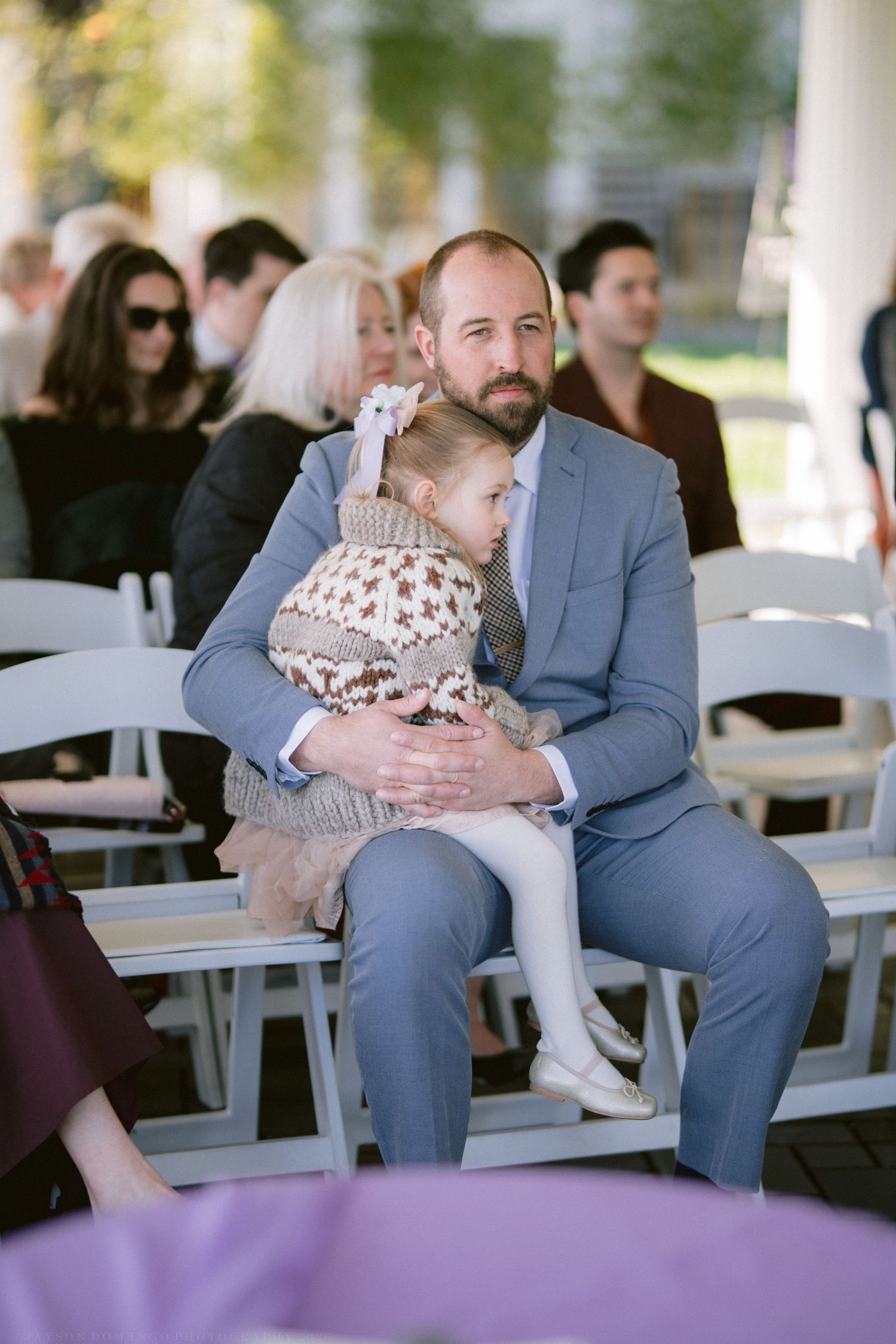 Father in gray suit holding young daughter during outdoor wedding ceremony.